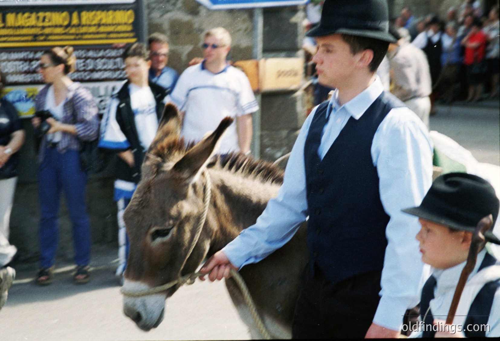 Vintage street scene featuring a man in a vintage vest and bowler hat leading a donkey, likely from a 1960s-1970s European festival or parade. Crowd in casual attire, including a child in traditional attire, stands behind. Signage in Italian ("Magazine A Repubbliche") suggests Italian location.