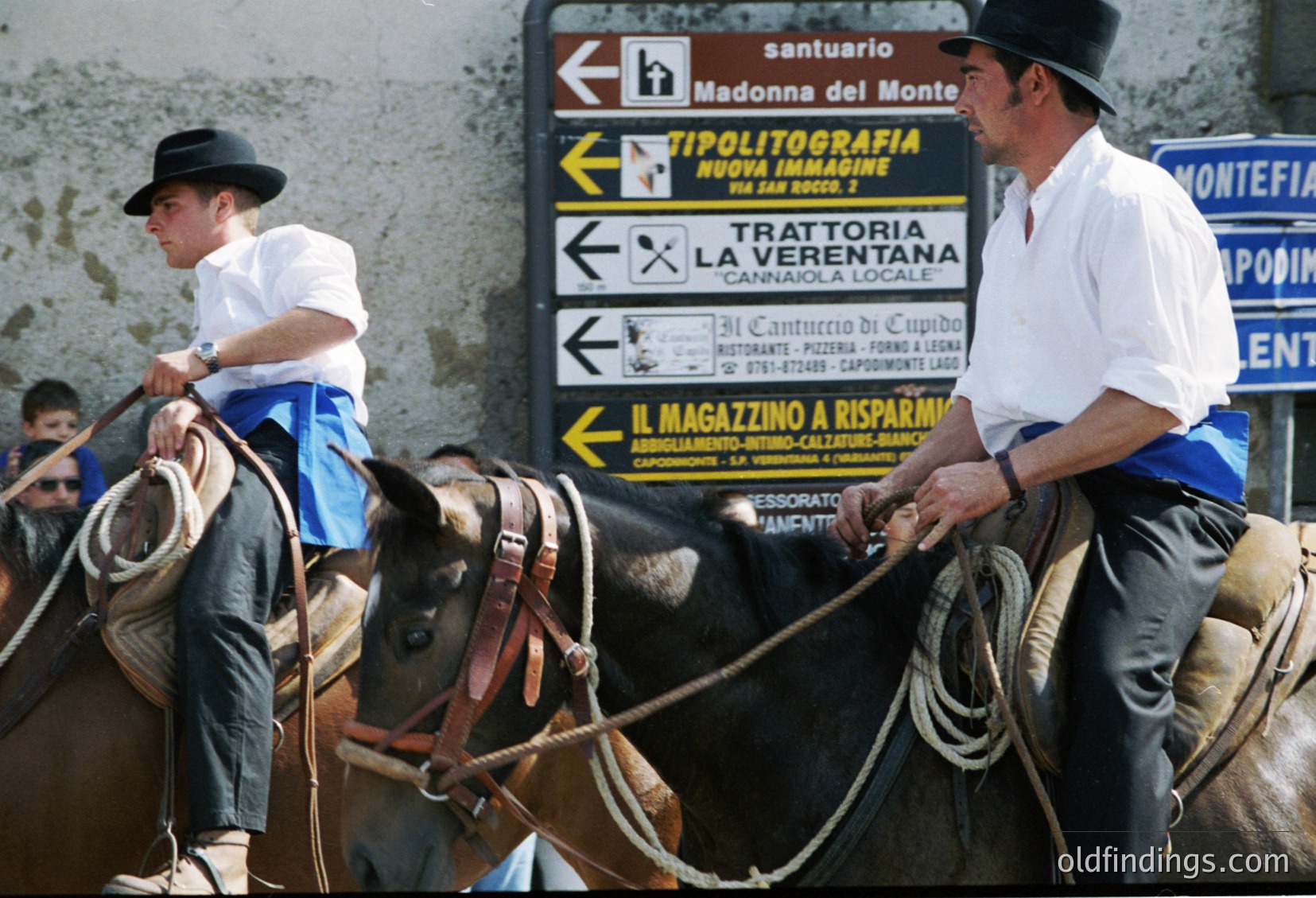 Two men in traditional Italian riding gear guide horses near a roadside signpost in . Signs indicate directions to local attractions like "Madonna del Monte" sanctuary and "La Verentana" trattoria, suggesting a rural or small-town setting. The attire and signage suggest a or timeframe. Ideal for cultural, historical, or travel-themed stock imagery.