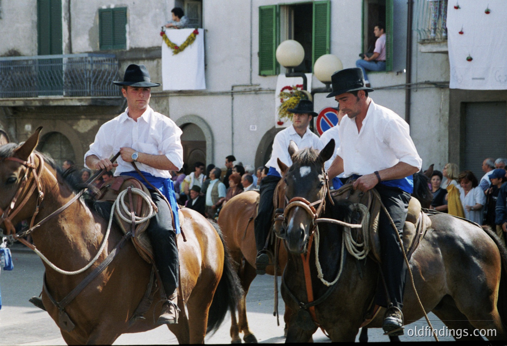 Riders in traditional white shirts, black hats, and blue trousers lead horses in a ceremonial procession through a European town square. Spectators line the background, observing from balconies and street level. The architecture features arched windows and pastel facades, suggesting a Mediterranean or Southern European setting. Likely from the mid-20th century, this image captures cultural heritage and community tradition.