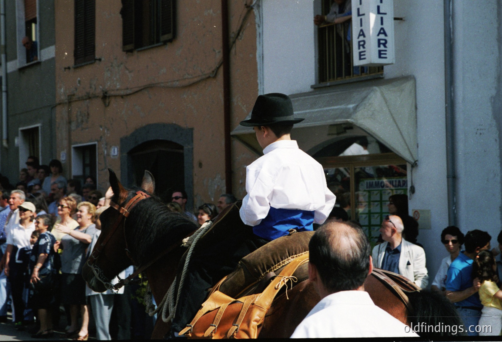 Traditional equestrian procession in a European village square, likely Italy’s Emilia-Romagna region. Riders in historical attire—white shirts, black hats, and saddled horses—participate in a festive event. Crowd of onlookers lines the cobblestone street. Signage in Italian suggests local shops.