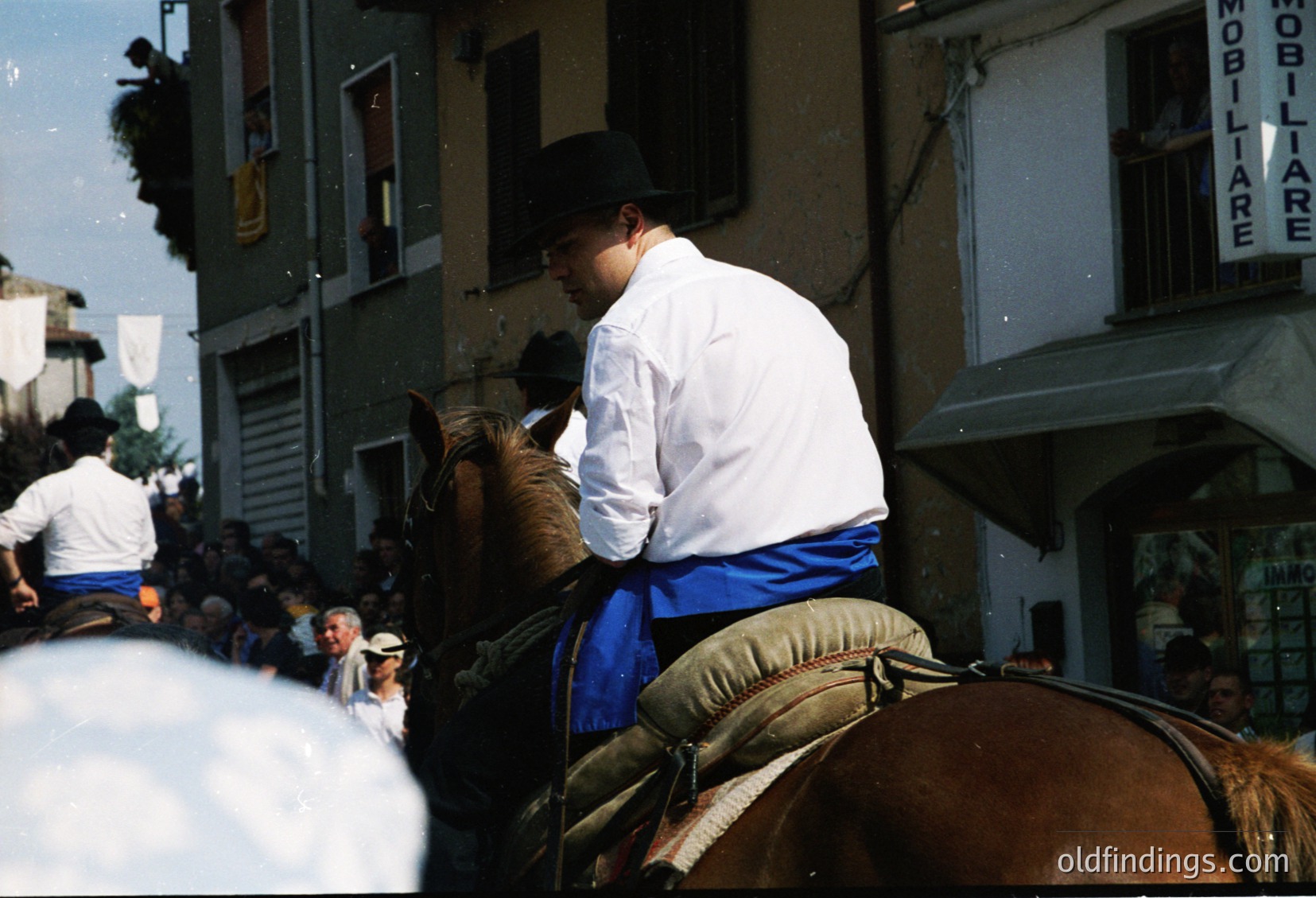 Rider in traditional alpine attire (white shirt, blue sash, black cap) leads a brown horse through a crowded street. Surrounding buildings feature Italian signage ("MOBILIARE") and balconies. Crowd in white attire suggests a cultural procession or festival. Likely