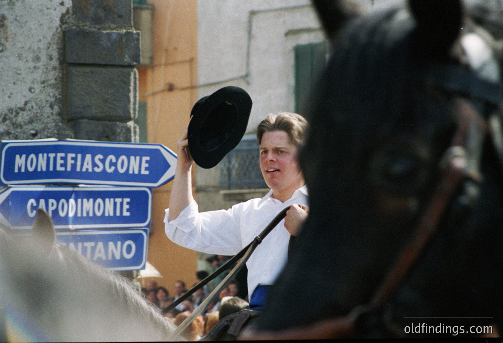 A man in a white shirt holds a fedora aloft near blue directional road signs ("Montefiascone," "Capodimonte," "Sant'Anna") in an Italian town square. Crowd and horse silhouettes frame the scene, suggesting a 1970s-80s public event or rally.