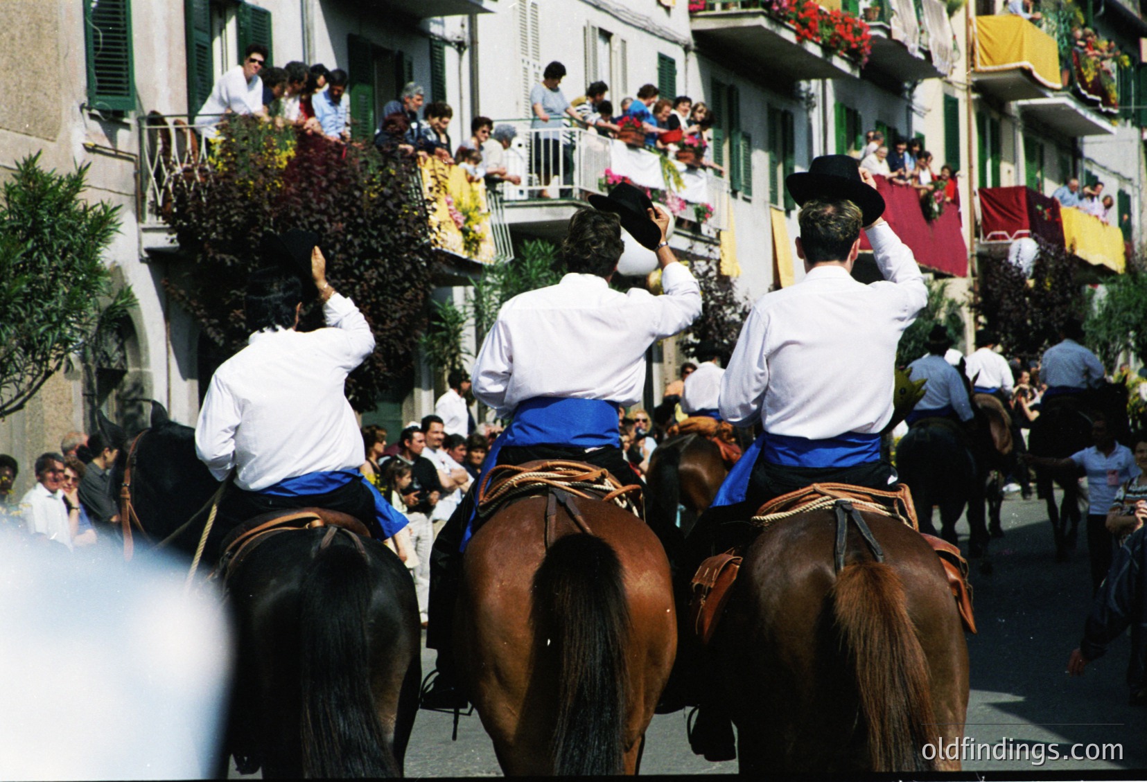 Three horsemen in traditional white jackets, blue sashes, and black hats carry floral arrangements during a festive procession. Crowds line balconies and streets, observing. Architectural details include wrought-iron railings and hanging flower baskets. Likely a Spanish cultural festival, possibly ín or similar.