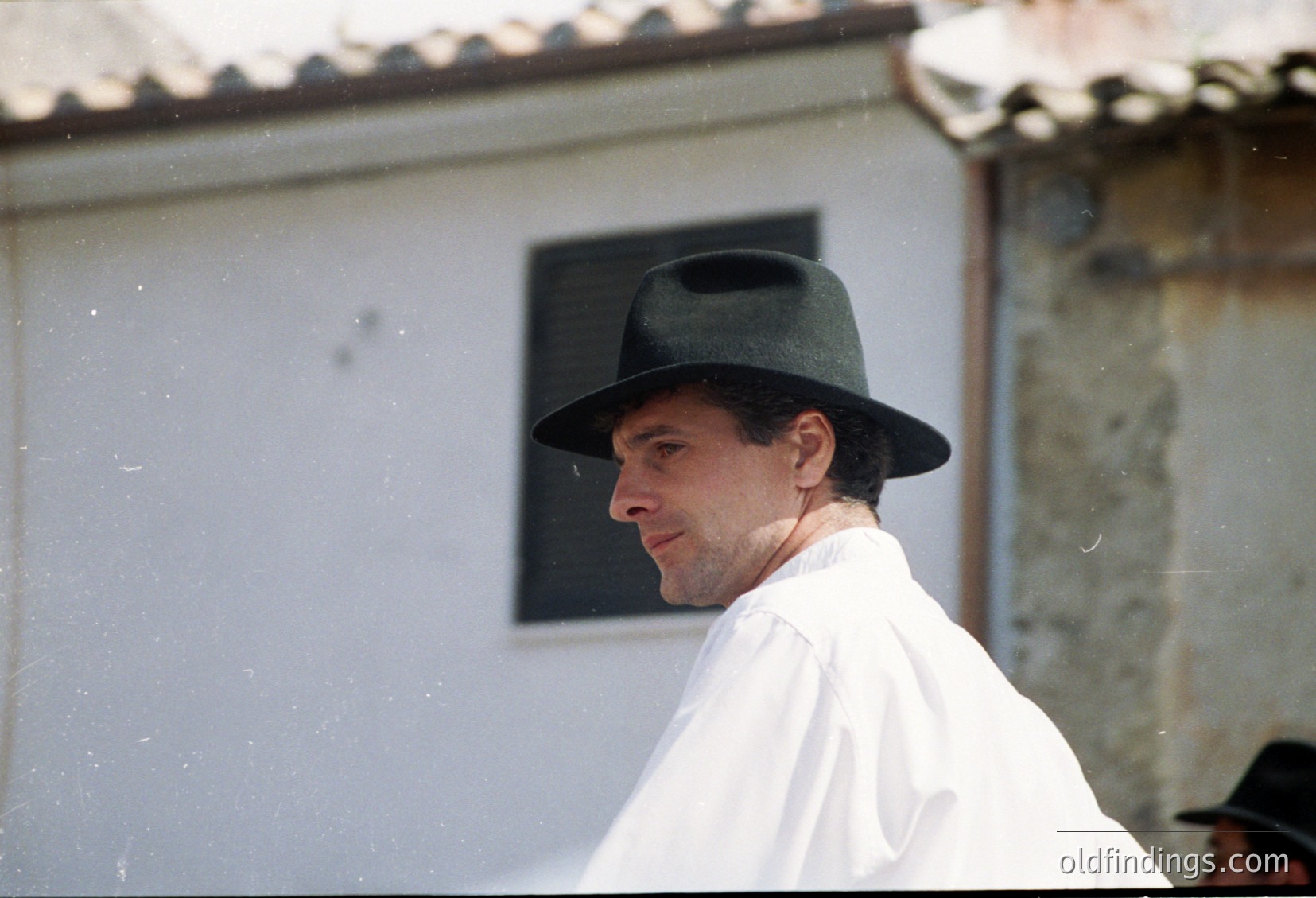 Portrait of a man in a wide-brimmed hat and light-colored robe, standing near a rustic stone building with a tiled roof. The architectural style suggests a Mediterranean or Southern European setting. Likely from the 1960s–1980s era.