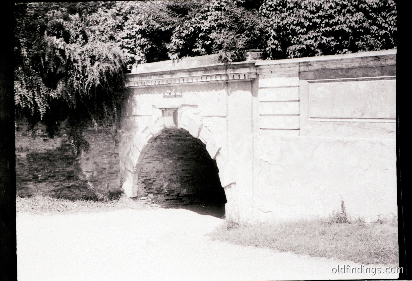 Historic stone archway with weathered brick tunnel entrance, flanked by high concrete walls. Likely part of a fortification or industrial site from the early-to-mid 20th century. Vegetation suggests outdoor setting.