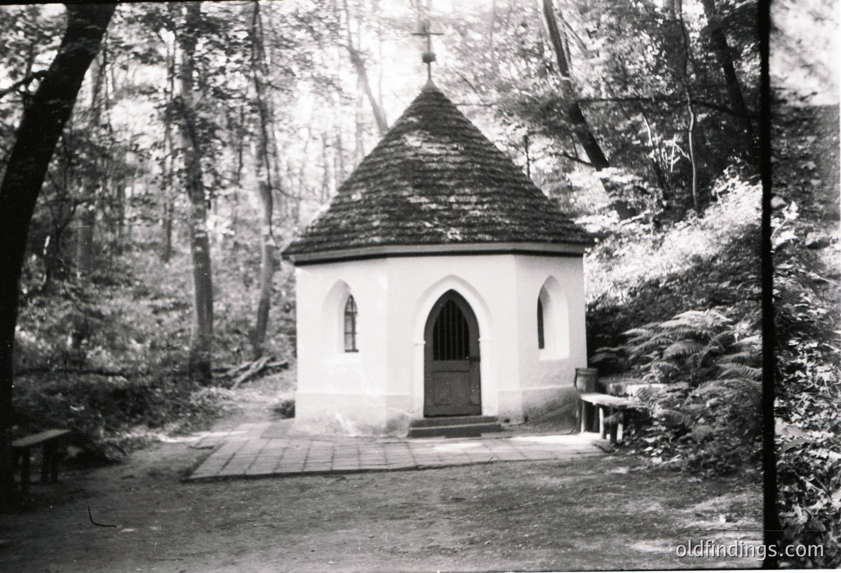 Isolated 19th-century alpine chapel with Gothic Revival arched doorway, conical slate roof, and cross-topped spire, nestled in dense forest. Stone steps lead to a small wooden bench beside a gravel path.