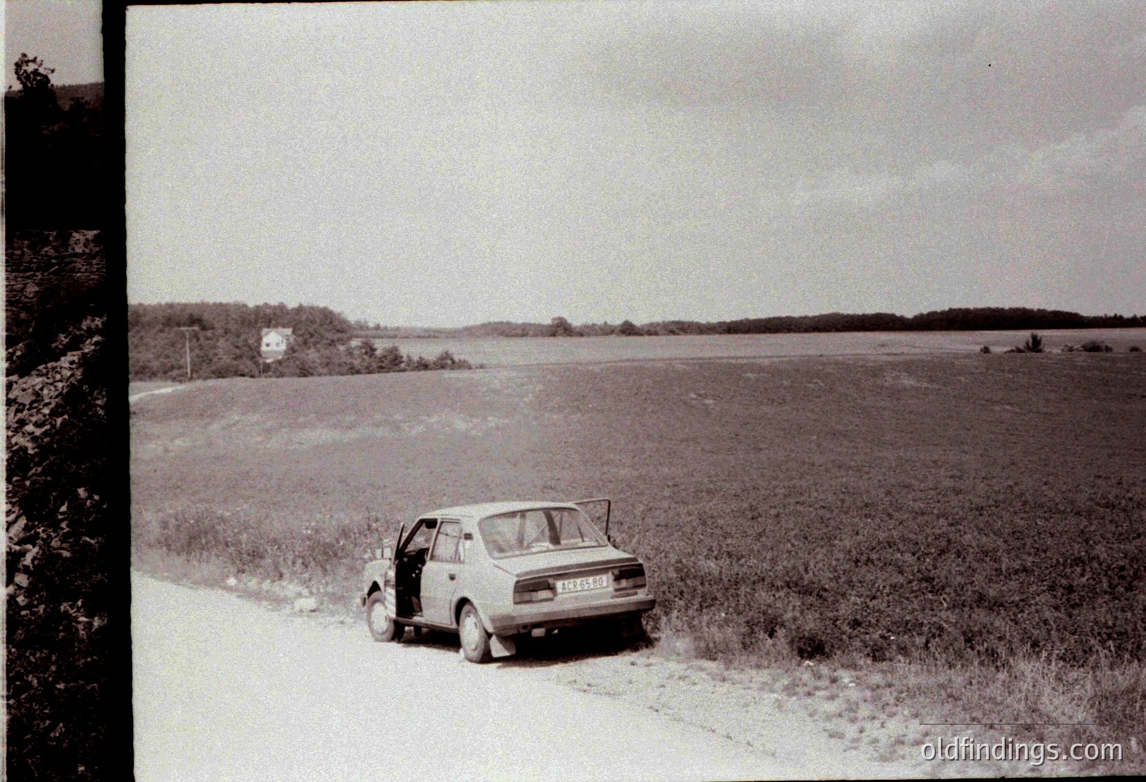 Vintage black-and-white shot of a compact sedan parked on a rural dirt road beside a vast, flat wetland. Distant tree line and single-story house visible across the water. Mid-20th century Eastern European countryside setting.
