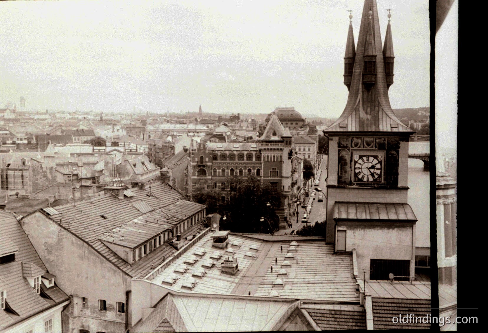 Aerial view of a European city square from the 1950s–1960s, showcasing a mix of historic and functional architecture. Prominent clock tower with ornate spire dominates right, while a grand, arched building with classical details anchors the center. Cobblestone plaza and low-rise structures reflect mid-century urban planning. River and distant skyline hint at a riverside location.