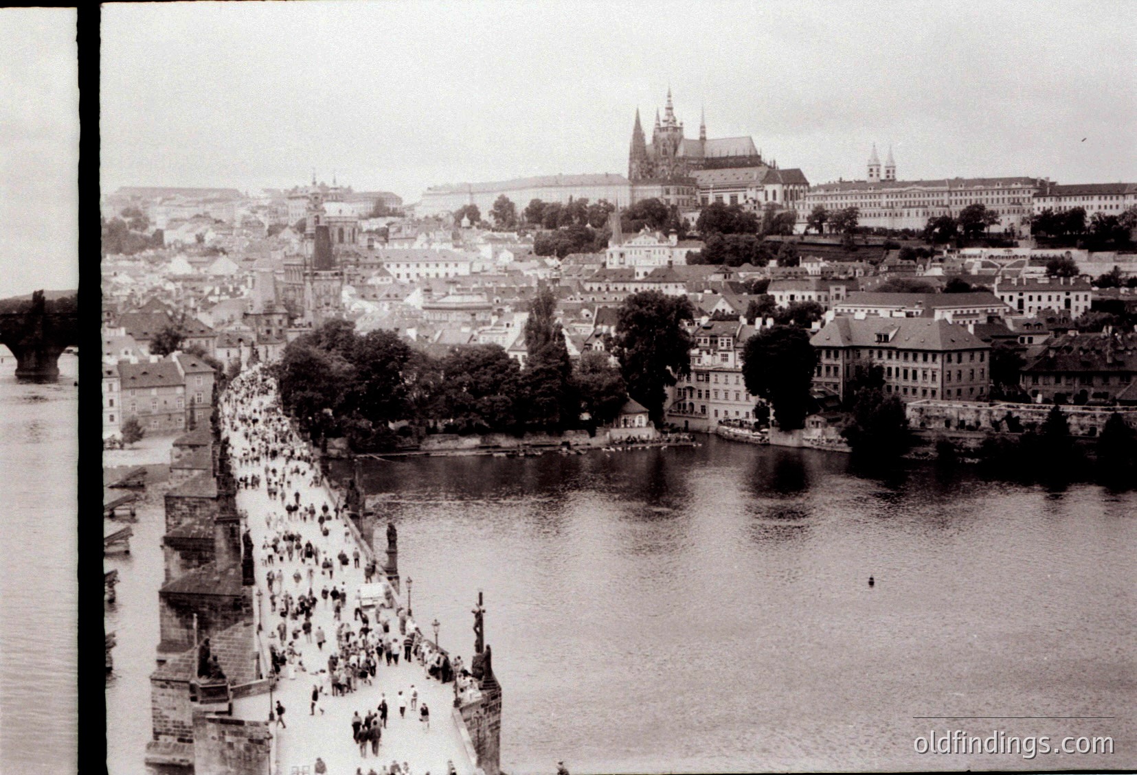 Vintage black-and-white aerial view of Prague’s Charles Bridge and Vltava River, crowded with pedestrians. Gothic spires of Prague Castle dominate the skyline, flanked by historic buildings. Mid-20th century (1950s–60s) urban landscape.