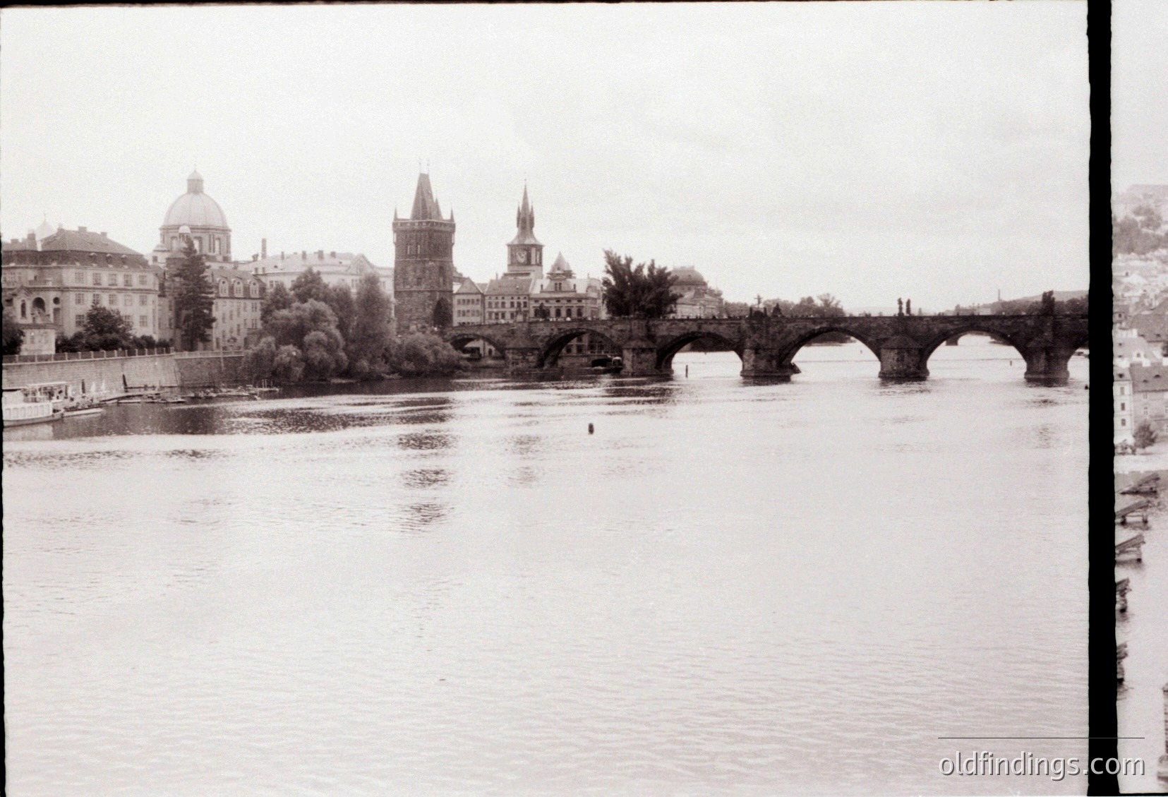 Vintage black-and-white view of Prague’s Charles Bridge spanning the Vltava River, flanked by historic towers and domes. Gothic-style architecture dominates the skyline, with a lone boat navigating the river. Early 20th-century urban landscape () captures the bridge’s iconic arches and riverbank details.