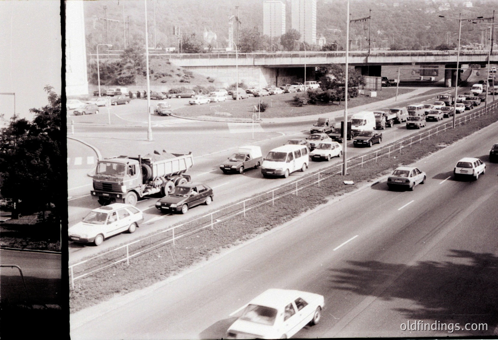 Mid-20th century urban highway jam with vintage vehicles, including a tow truck assisting a disabled car. Concrete barriers and elevated highway in background suggest mid-century infrastructure. Traffic congestion highlights early highway challenges.