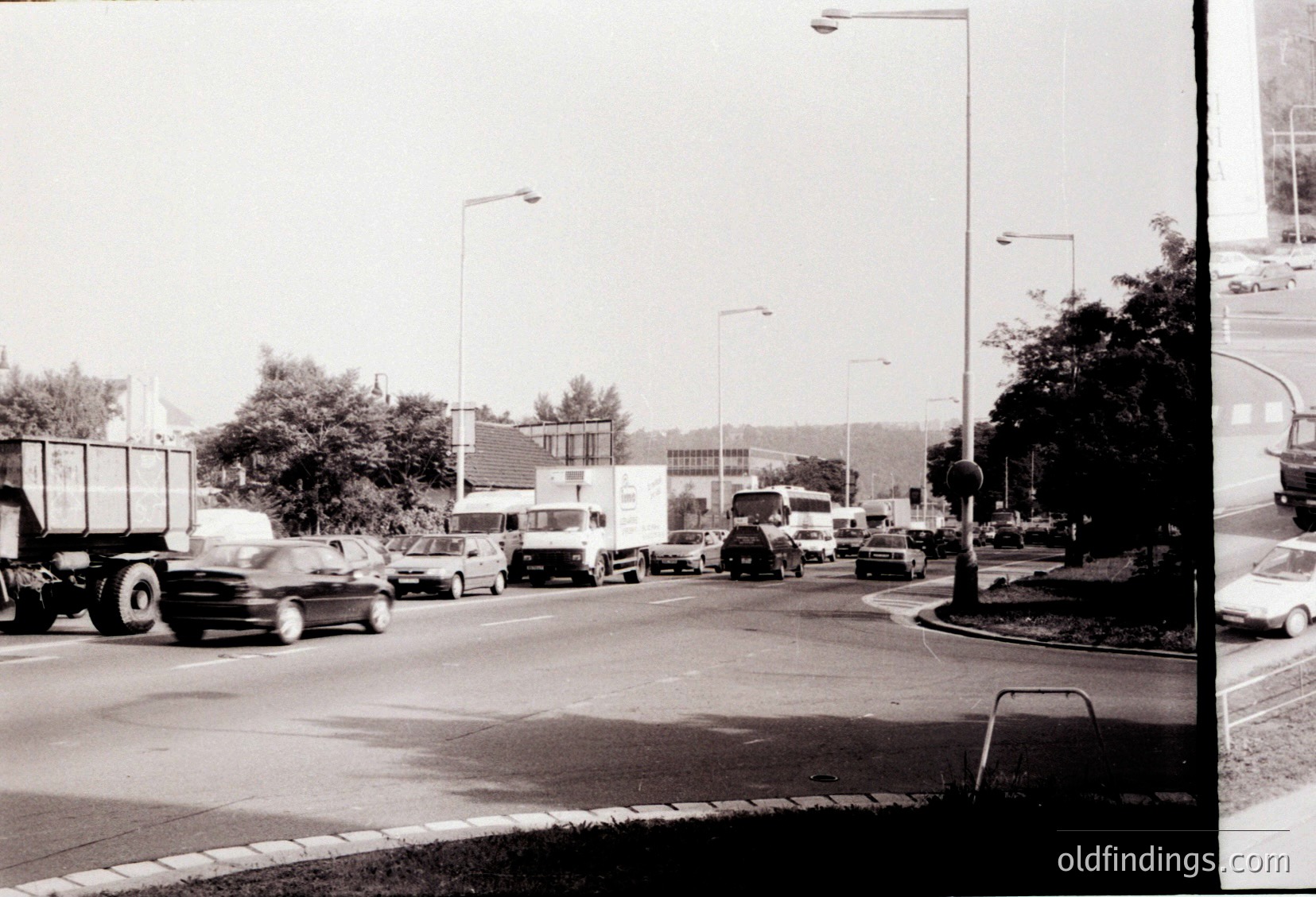 Urban traffic scene featuring mid-20th century vehicles, including a truck and sedans, navigating a roundabout. Buildings and streetlights suggest a planned city layout. Likely or era, possibly in a Western European or North American city.
