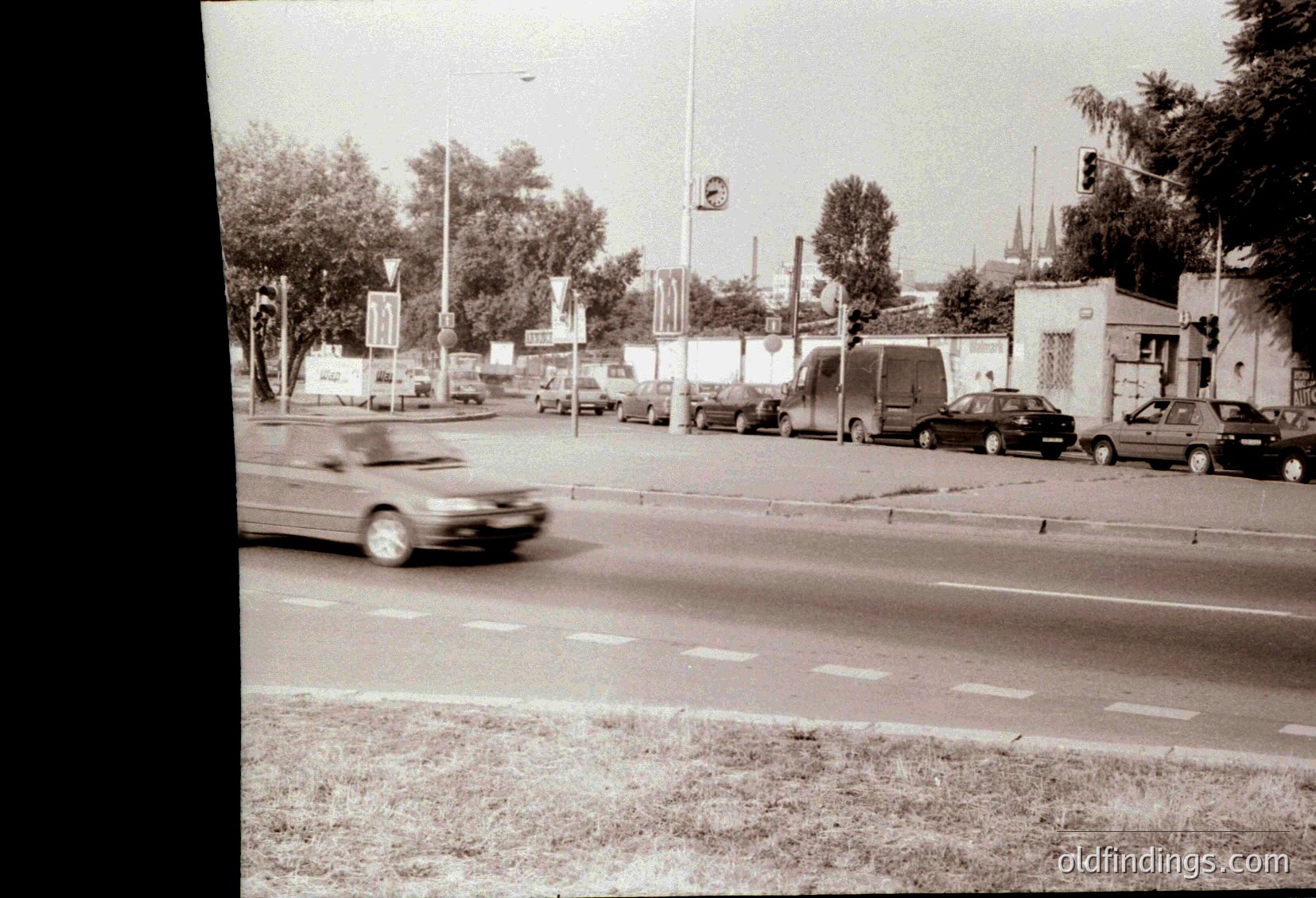 Urban street scene with motion blur of a passing car, lined with parked vehicles and greenery. Mid-20th century architecture and signage suggest a European city. Busy roadside with trees, parked cars, and a visible speed limit sign.