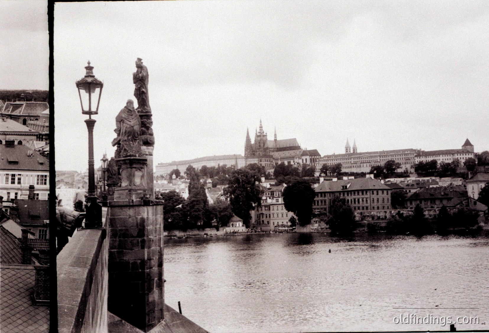 Black-and-white view of Prague’s **Charles Bridge** with ornate stone statues flanking the railing. Gothic spires of **Prague Castle** and **St. Vitus Cathedral** rise in the background across the Vltava River. Mid-20th century (1950s–1960s) urban landscape, showcasing historic European architecture.