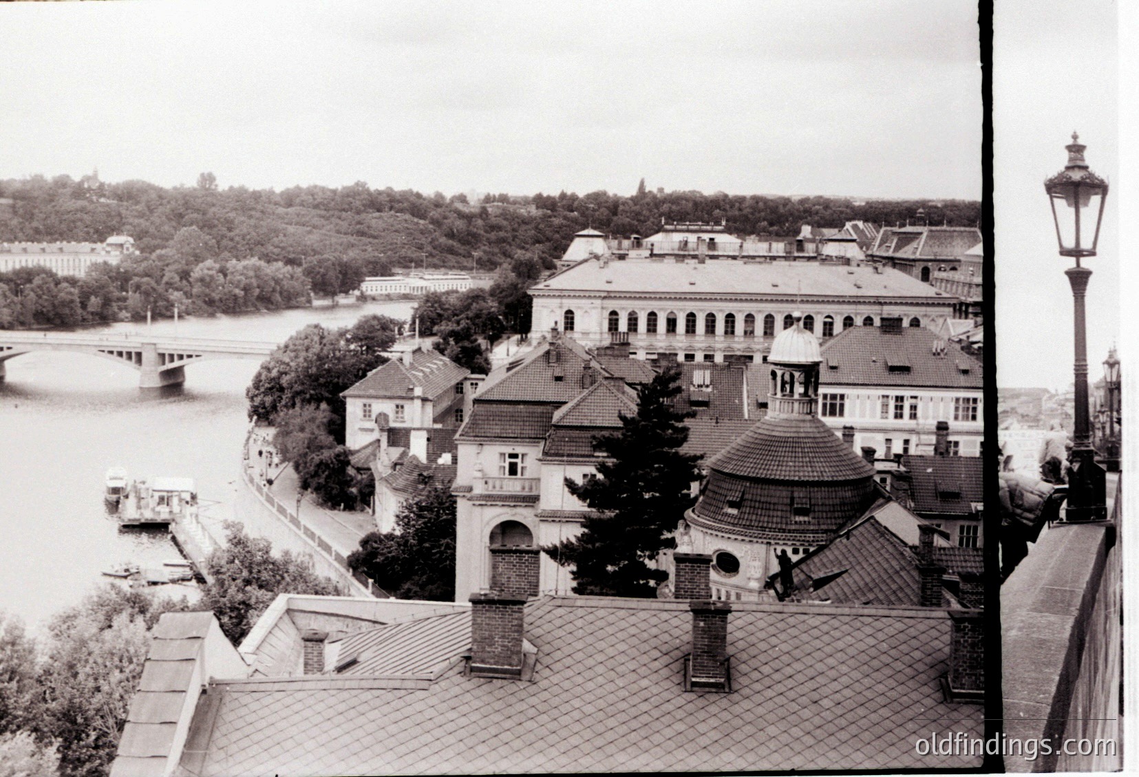 Black-and-white urban riverside scene featuring early 20th-century European architecture. Prominent domed building with red-tiled roof and arched windows, flanked by classical-style structures along a riverbank. Bridge and lush greenery in background. Likely .