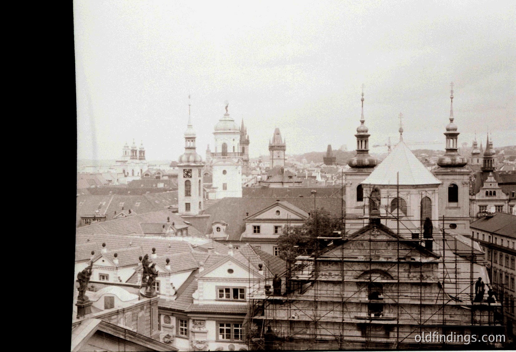 Early 20th-century European cityscape featuring Baroque-style domes and spires. Prominent church with scaffolding suggests restoration. Low-angle view captures rooftops and distant skyline under overcast skies. Likely Eastern European due to architectural style.