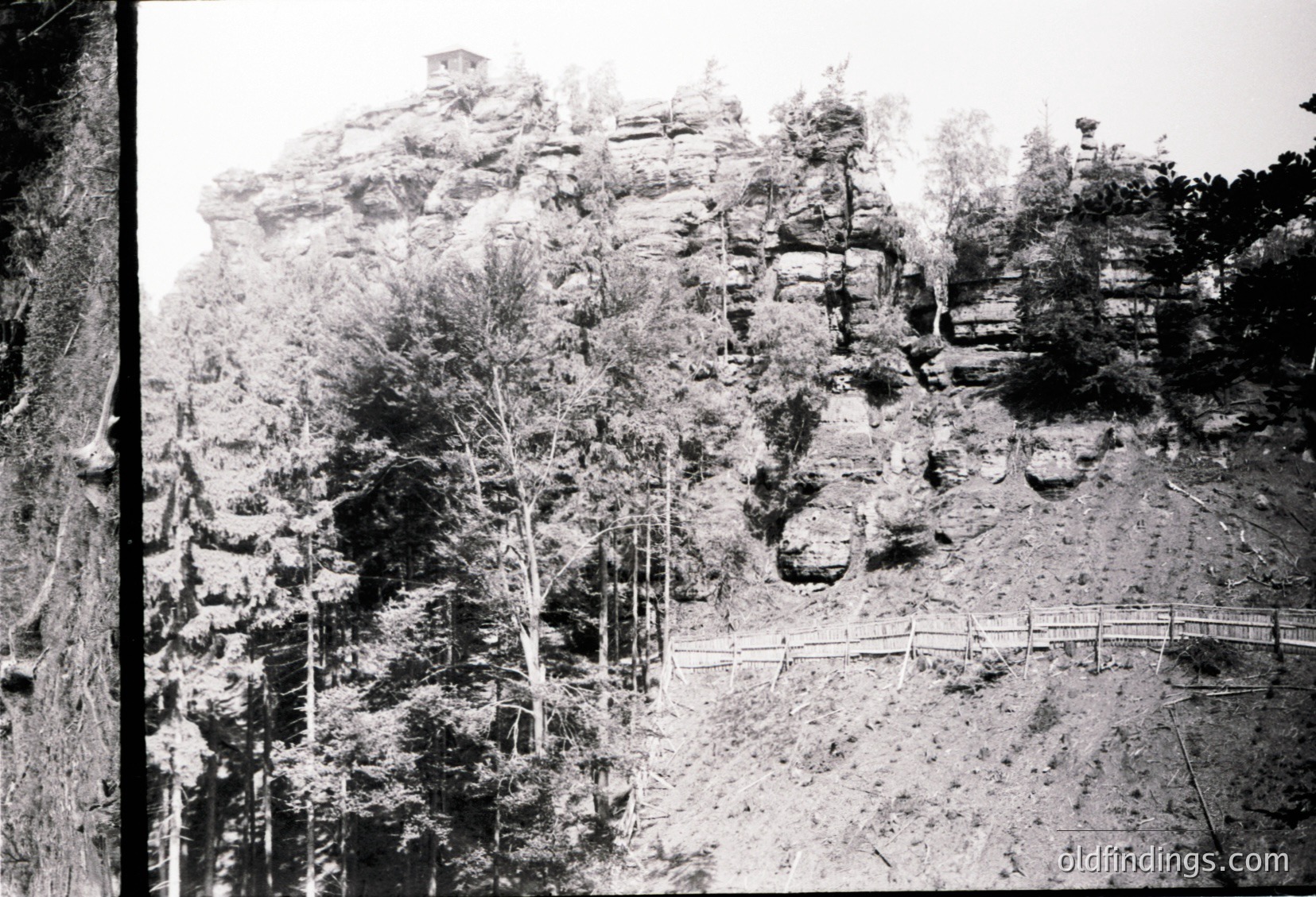 Early 20th-century black-and-white landscape of rugged rock formations and forested slopes. A wooden fence traces a path along the base, leading into the dense foliage. Likely Eastern European due to terrain and vegetation.