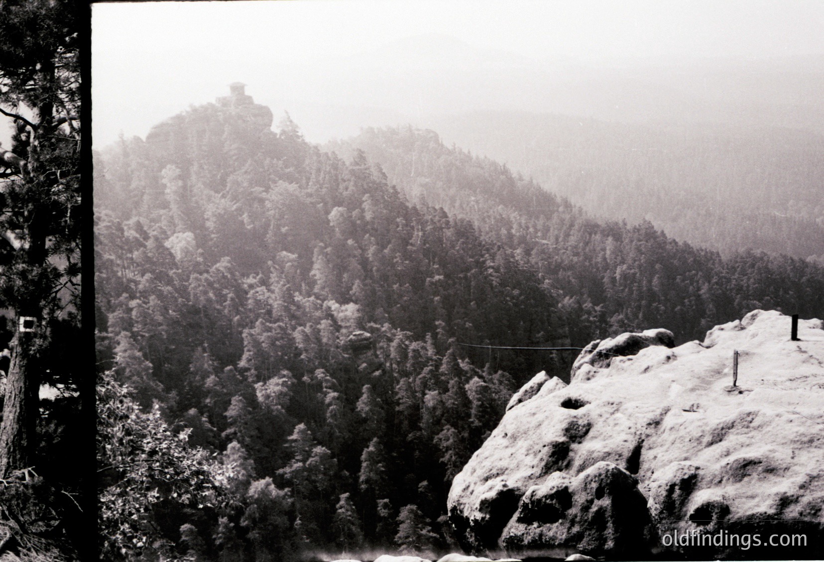 Snow-covered rocky outcrop with dense forest below, misty alpine backdrop. Black-and-white vintage photograph, likely 20th century. Dramatic winter landscape with rugged terrain and dense coniferous trees.