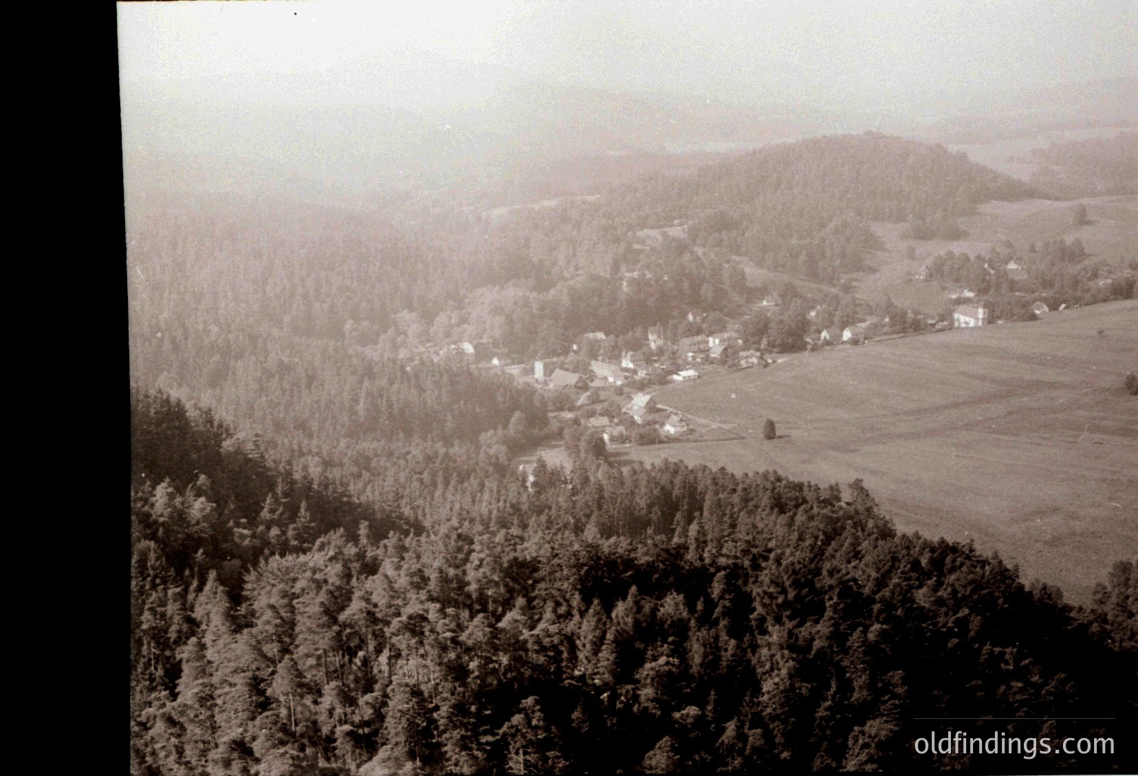 Aerial vintage photograph of a rural valley, likely 1940s–1960s. Dense forest frames the lower left, while open fields and scattered farmhouses occupy the valley floor. Mist lingers over rolling hills and distant treeline, suggesting early morning or autumn.