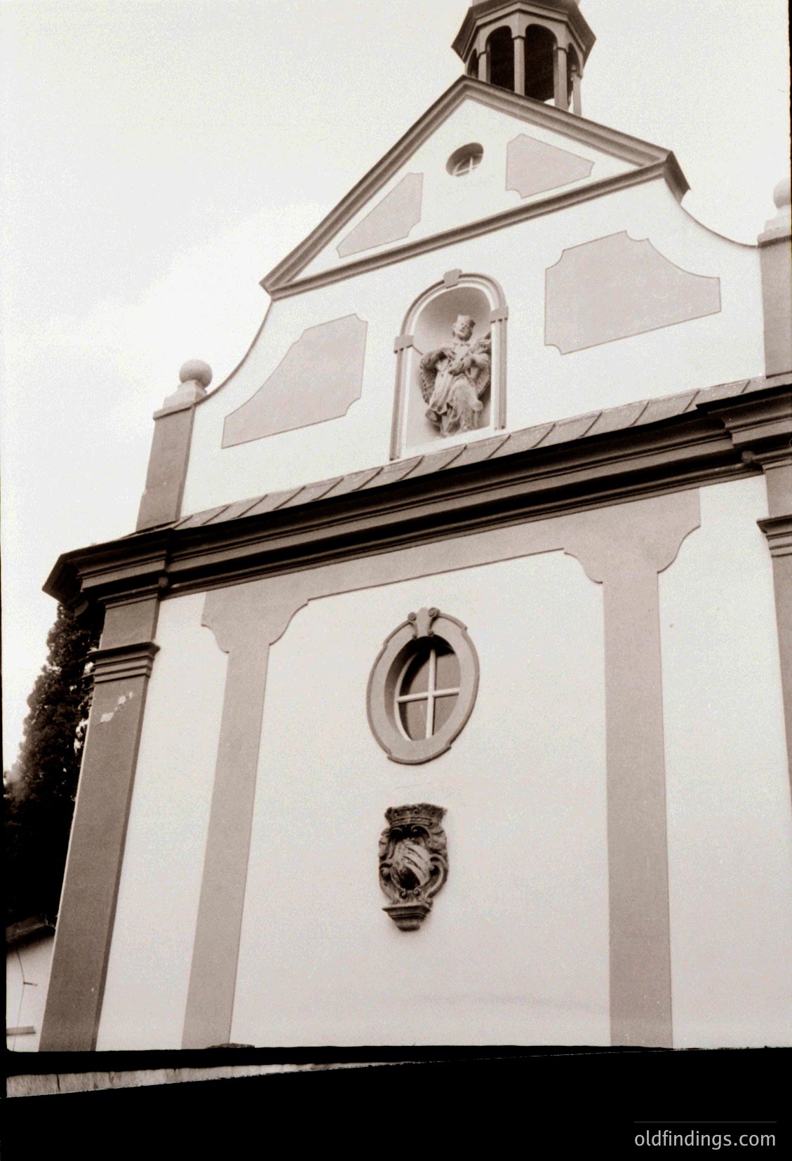 Neoclassical church façade featuring a symmetrical design with a central pediment and bell tower. The white plaster walls display a round window with a cross motif and an ornate coat of arms below. Religious relief sculpture above the entrance depicts a saint. Likely European, 18th–19th century.
