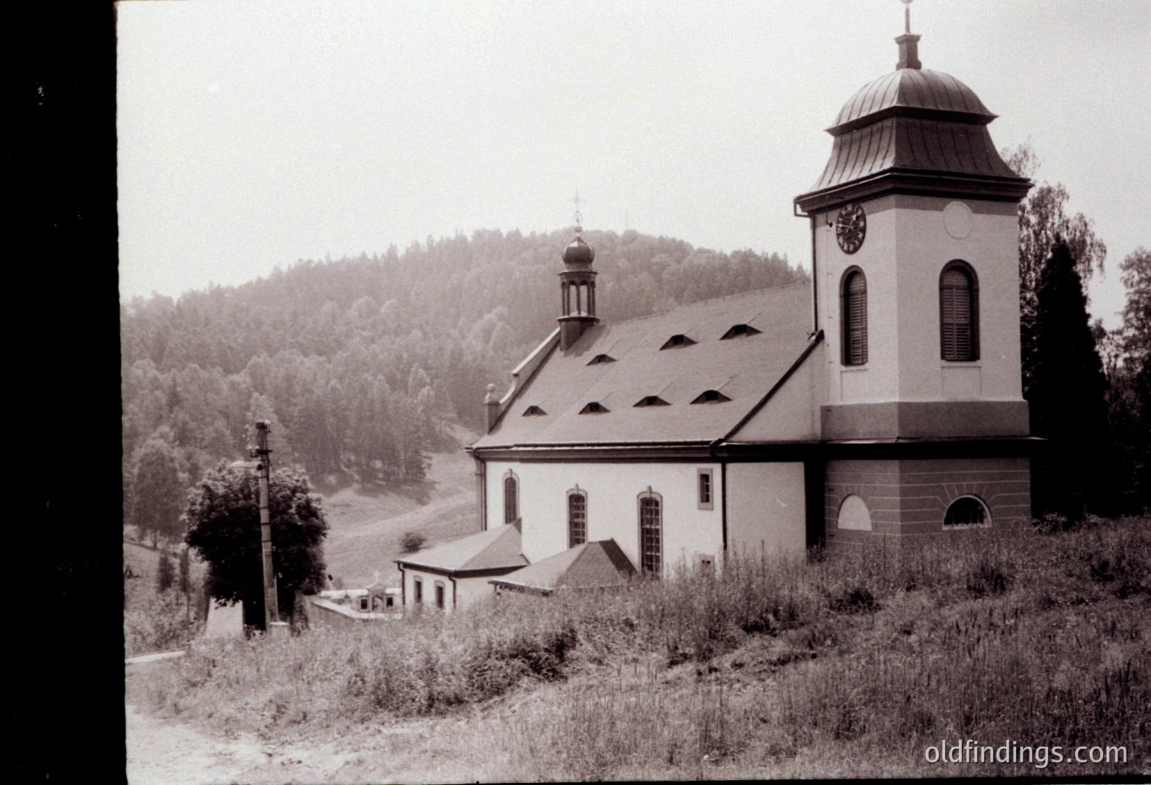 Mid-20th century church with distinct bell tower and clock, set against forested hills. Brick construction with arched windows and gabled roof. Rural landscape with overgrown grass and utility poles. Likely Eastern European architecture.