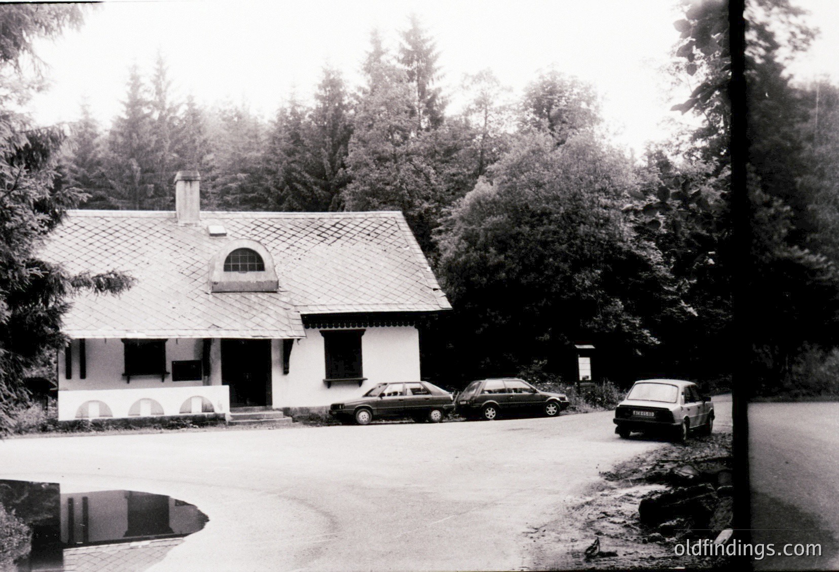 Single-story mid-century suburban home with curved roof edges, arched windows, and a tiled facade. Three vintage cars (1960s-70s) parked in a gravel driveway surrounded by dense evergreen forest. Classic post-war residential architecture.