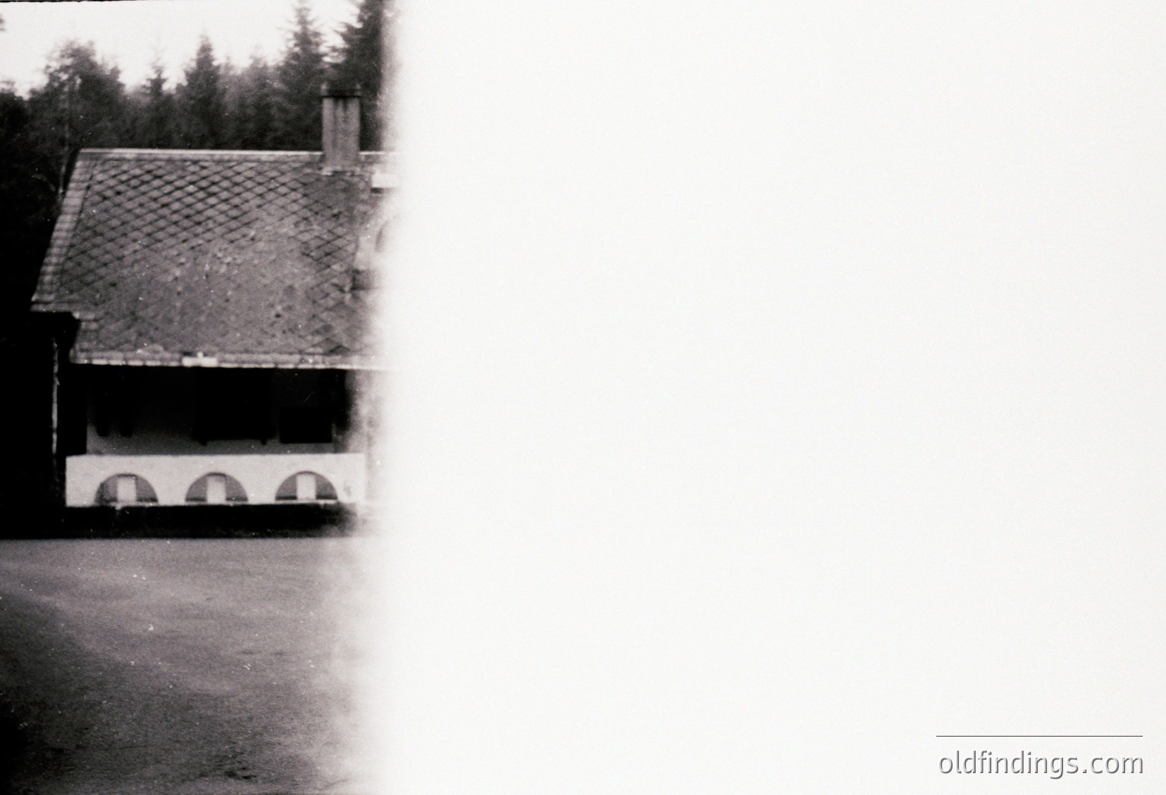 Vintage black-and-white shot of a rustic stone house with a tiled gable roof and arched basement windows. Dense forest backdrop suggests rural isolation. Likely mid-20th century European countryside architecture.