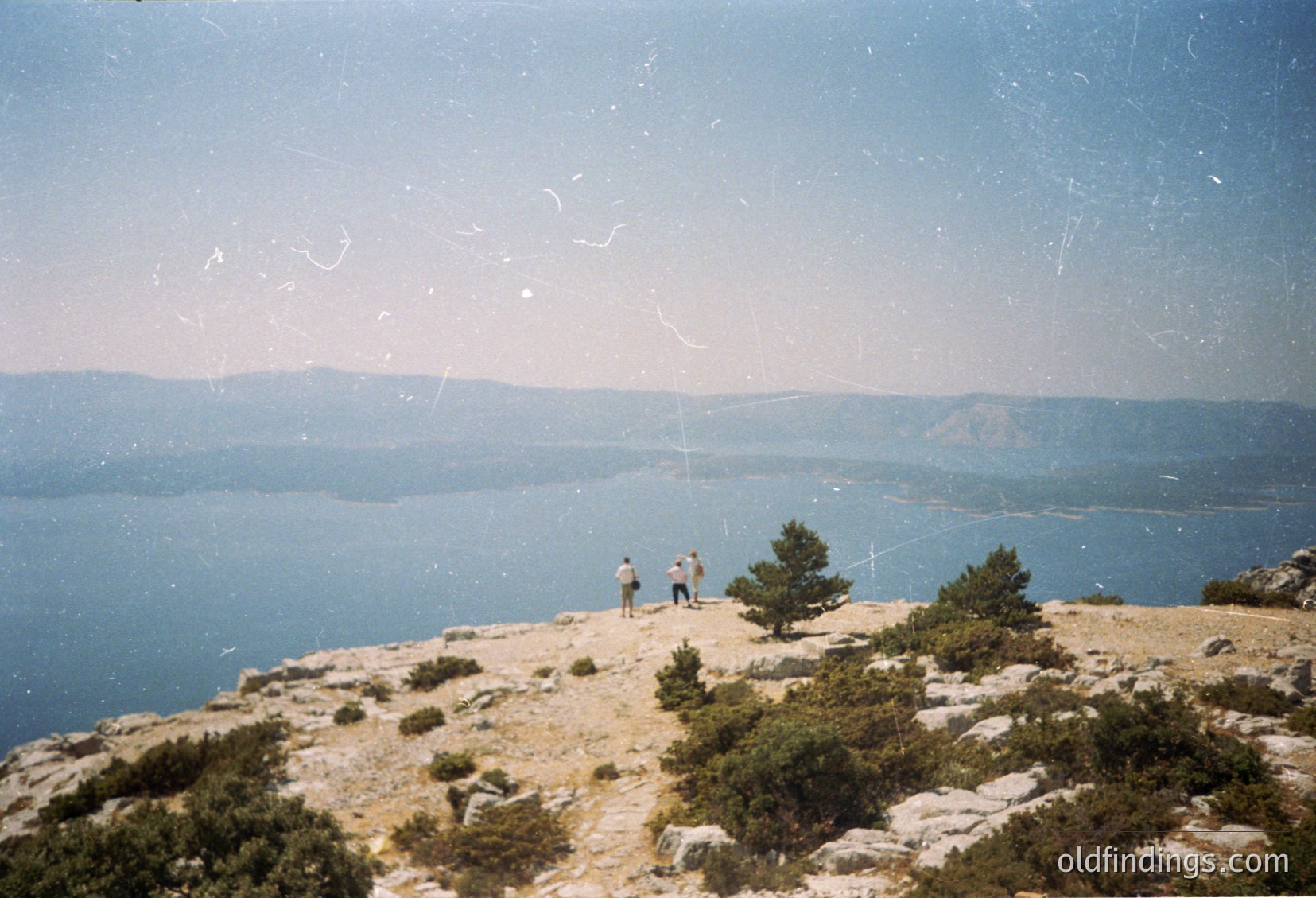 Vintage coastal landscape shot with three figures atop rocky terrain overlooking a deep blue sea and distant landmass. Minimal vegetation, sparse shrubs, and clear horizon line. Likely Mediterranean or similar climate.