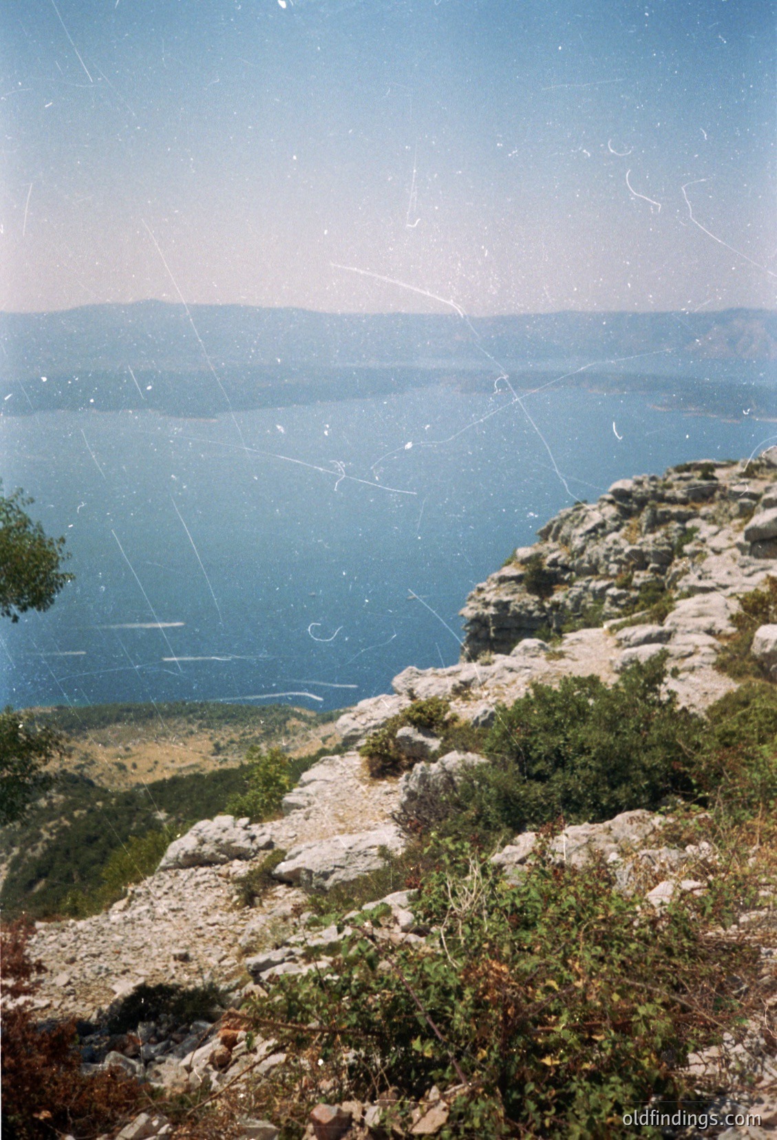 Vintage photograph showing a rugged coastal cliffside with sparse vegetation and a deep blue sea extending to distant landforms. The rocky terrain contrasts with the serene horizon. Likely captured on analog film with visible scratches and grain.