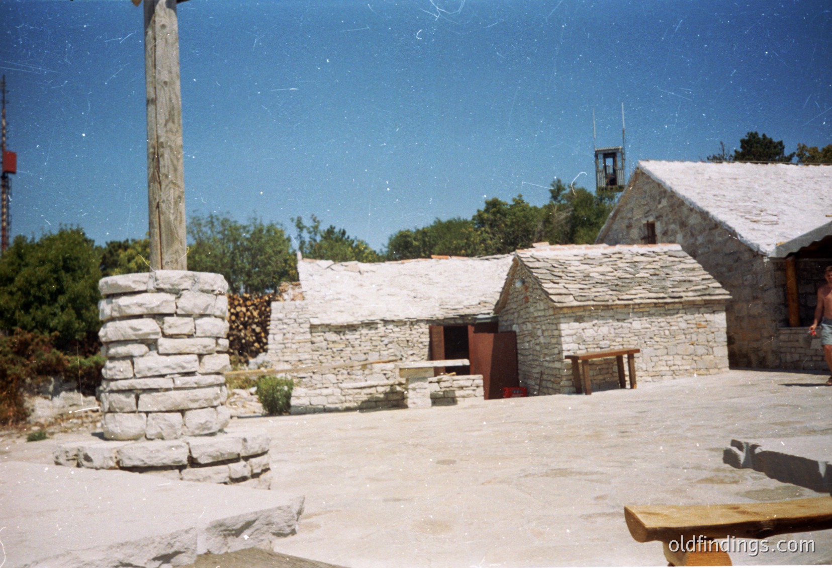 Rustic stone courtyard with traditional rural architecture—stone-built huts with pitched roofs, a wooden cross, and a stone pedestal. Likely Eastern European, possibly or , with -1980s aesthetic. Ideal for historical research or folk-inspired design references.