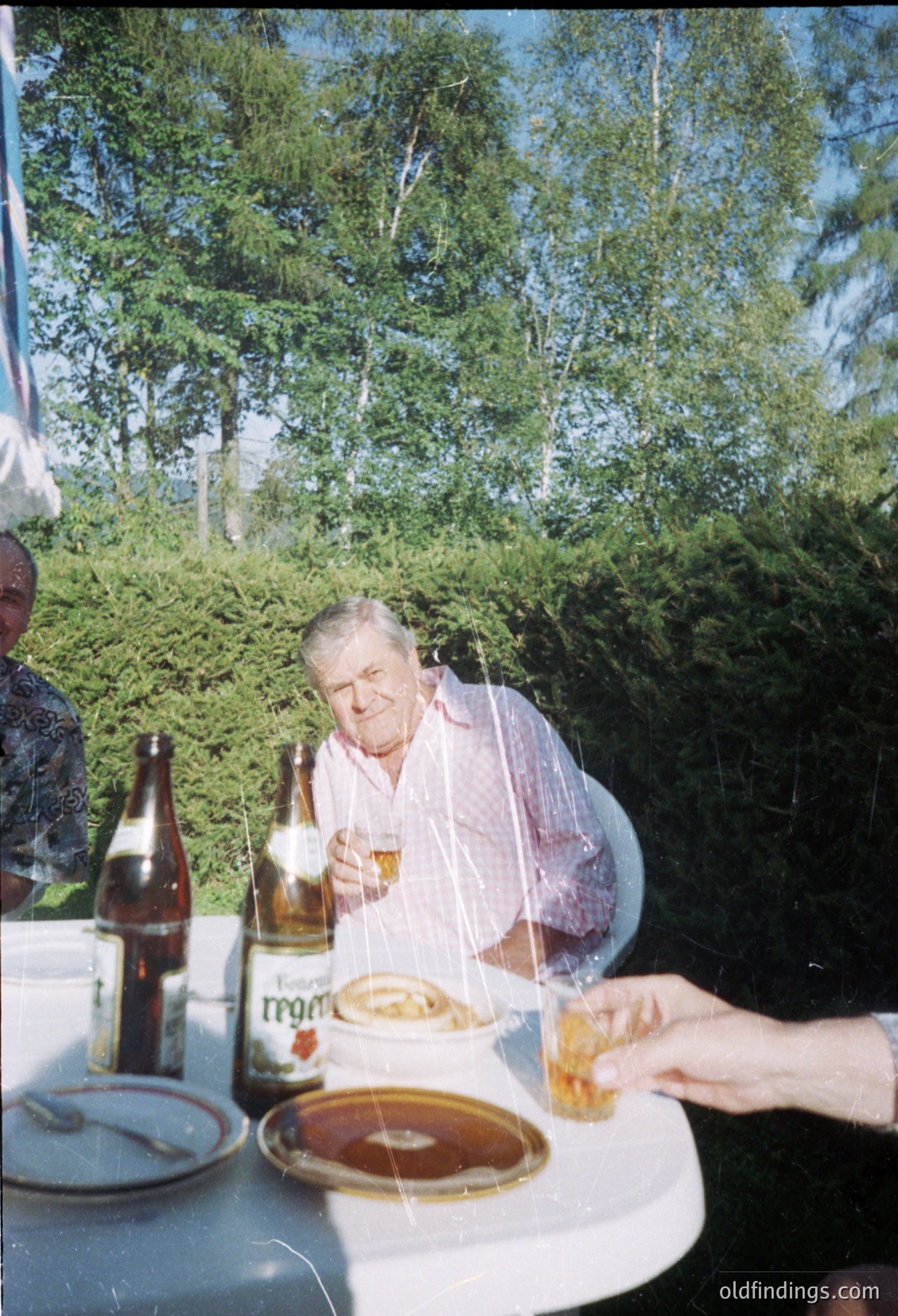 Mid-century outdoor dining scene featuring an older man in a light-colored shirt, seated at a round table with a bottle of *Regent* beer and a plate of food. Another hand reaches across to serve or share. Lush greenery and trees in the background suggest a garden or patio setting, likely in a European country. Vintage aesthetic with warm lighting.