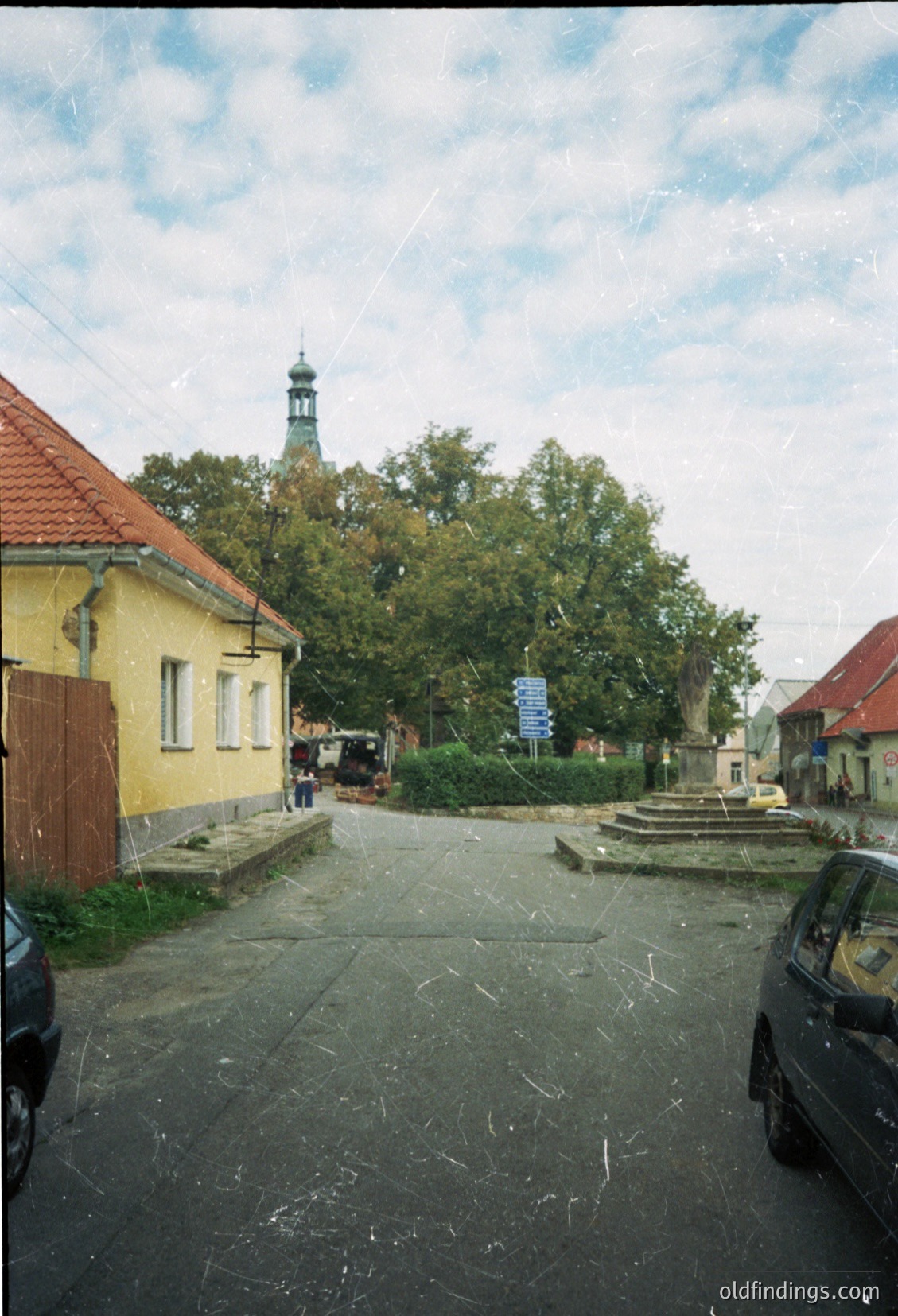 Vintage street scene featuring Soviet-era architecture: yellow-painted brick house with red-tiled roof, cobblestone courtyard, and a lone tree framing a church tower in background. Two parked cars suggest mid-20th century setting.