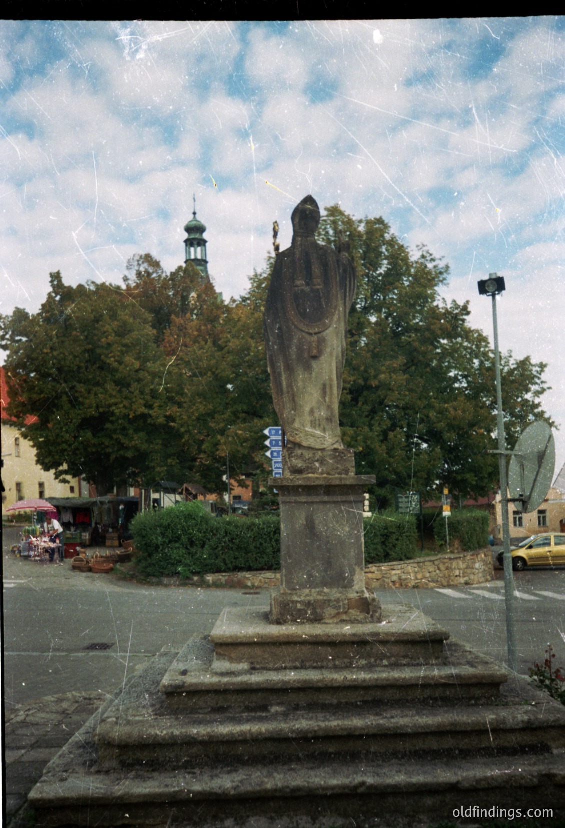 Stone statue of a robed figure holding a cross, mounted on a pedestal in an urban square. Surrounding elements include directional signs, trees, and a church steeple in the background. Likely European, mid-20th century.