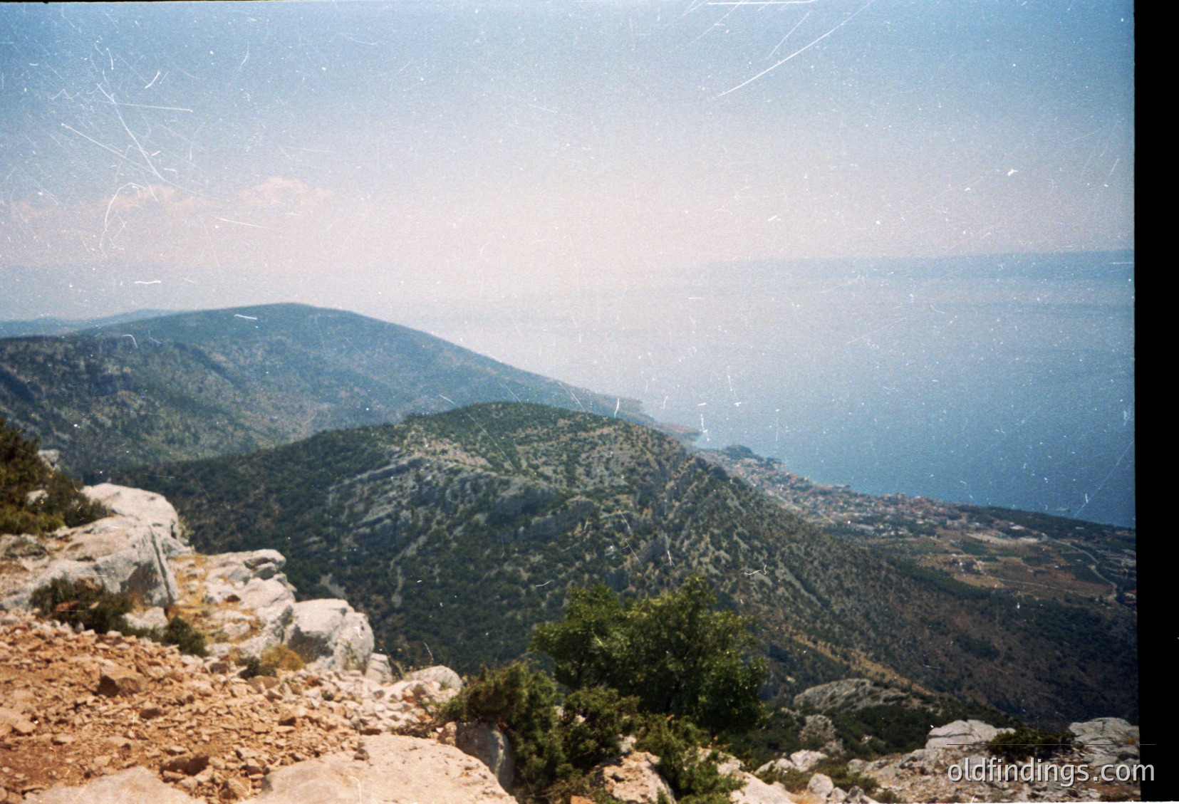 Vintage panoramic shot of rugged coastal mountains meeting the sea, likely Mediterranean. Dense greenery contrasts with rocky terrain and distant urban sprawl. Film grain and slight blur suggest mid-20th century photography (). Ideal for travel inspiration or historical geography studies.