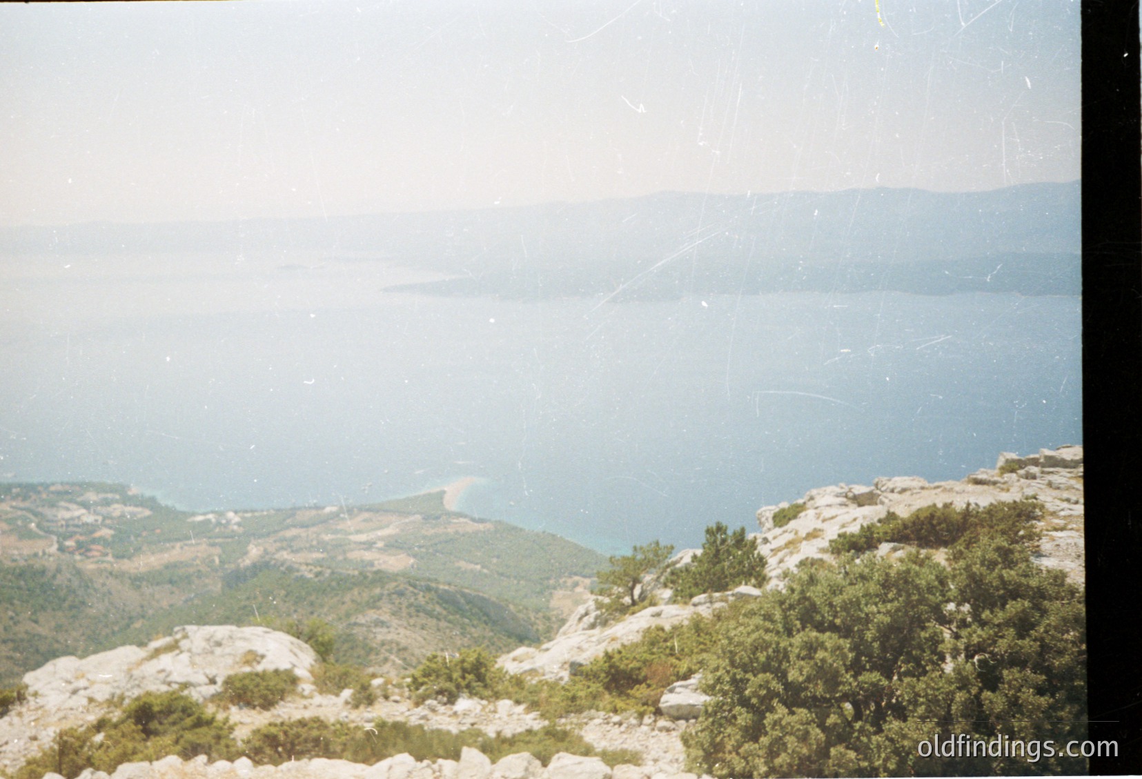 Vintage coastal landscape shot from elevated rocky terrain, showcasing a serene sea meeting distant land. Vegetation includes sparse trees and shrubs, with a hazy horizon blending sky and water. Likely Mediterranean or Black Sea region.
