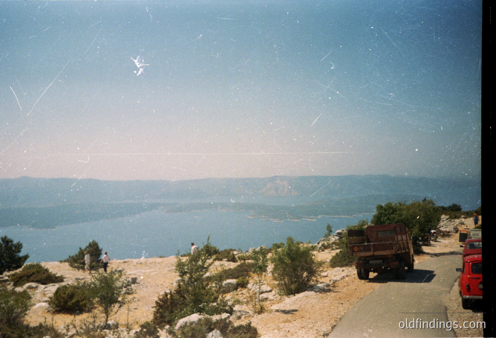 Vintage coastal roadside scene with rugged terrain and deep blue sea. A lone red truck parked beside a winding asphalt path, bordered by sparse vegetation and rocky outcrops. Distant figures near the water’s edge suggest a quiet seaside spot. Likely Mediterranean or Black Sea region, 1970s–1990s.