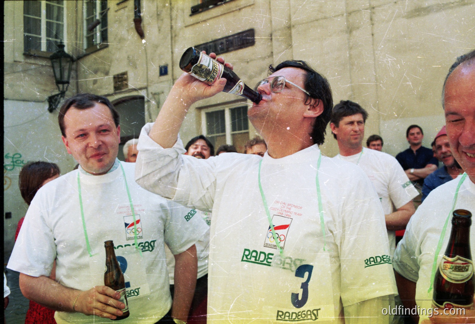 Celebratory moment at the 1980 Moscow Olympics, featuring two men in matching white "Rade Gast" event shirts drinking beer. The man in the center, wearing bib , chugs from a bottle while splashing others. Urban setting with Soviet-era architecture in the background.