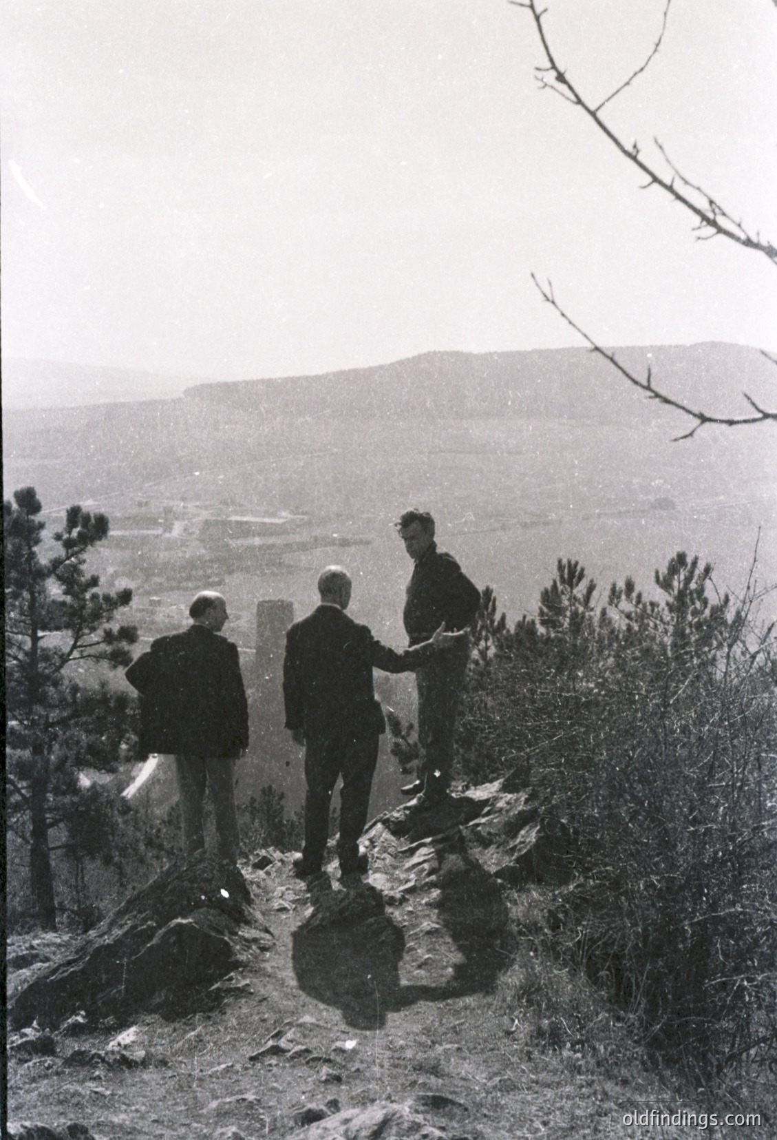 Three men in formal attire pose atop rugged terrain, overlooking a valley with distant hills. Mid-20th century black-and-white photo, likely 1950s–1960s. Formalwear suggests a ceremonial or official gathering.