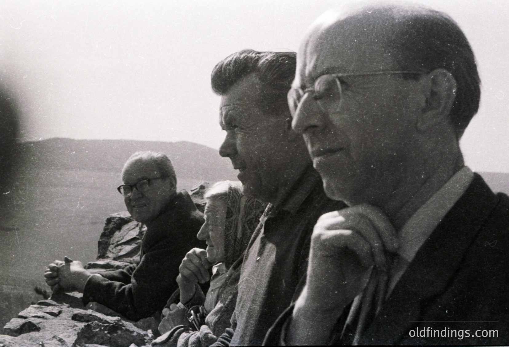 Three men in mid-20th-century attire pose outdoors on rocky terrain, likely a hillside or mountain. The central figure wears a patterned jacket and glasses, while the others in dark suits and glasses appear in profile. Black-and-white format suggests 1950s–1960s era.
