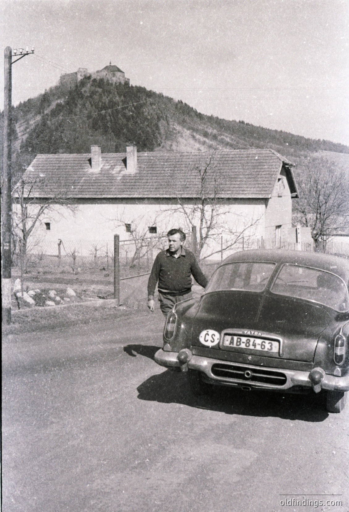 Mid-20th century black-and-white photo featuring a man in a dark jacket and cap standing beside a vintage **Škoda AB-84-63** car (1960s Czechoslovakia). Rustic stone hilltop fortress looms in background, with a single-story white farmhouse and barren trees in foreground. Rural European countryside setting. #ŠkodaAB84
