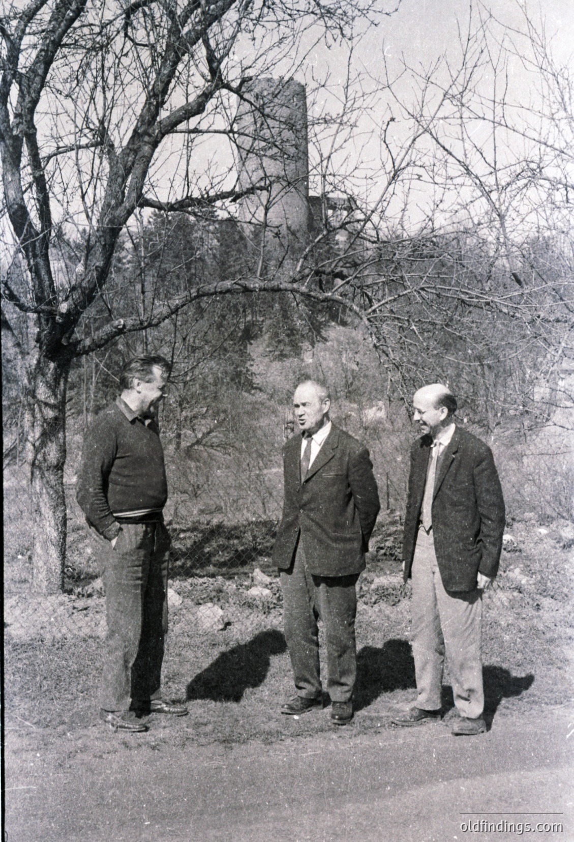 Three men in mid-20th-century attire stand near a partially demolished stone structure, likely a historic ruin. The man on the left wears a sweater and jeans; the center figure sports a suit and tie; the rightmost man wears a light shirt and tie. Leafless trees suggest late autumn/winter.