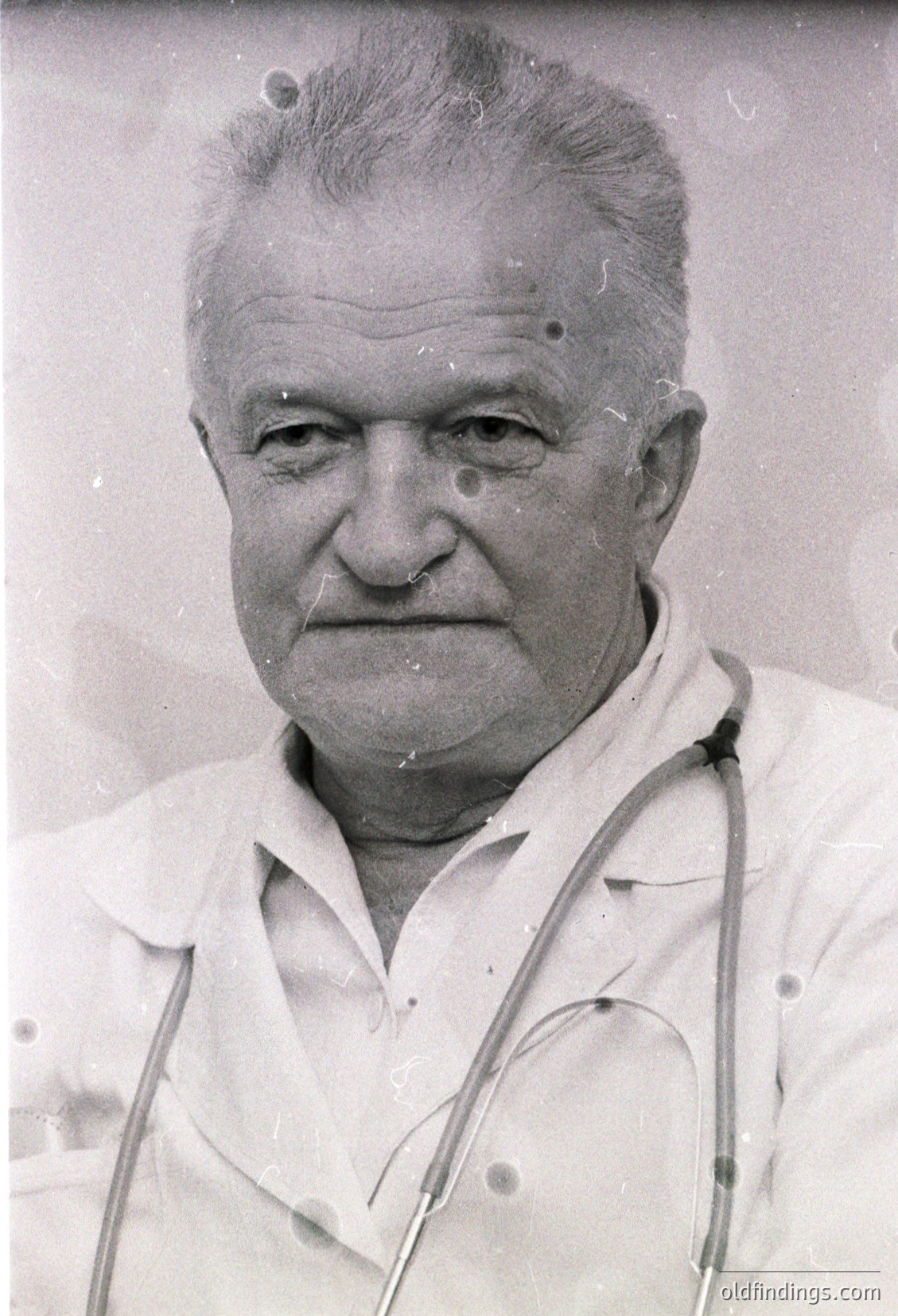 Portrait of an elderly man in a vintage stethoscope draped over his shoulders, suggesting a medical profession. The grainy, monochrome style and slight blur indicate a mid-20th century photo, likely 1940s–1960s. Neutral expression and classic hairstyle reflect historical fashion.
