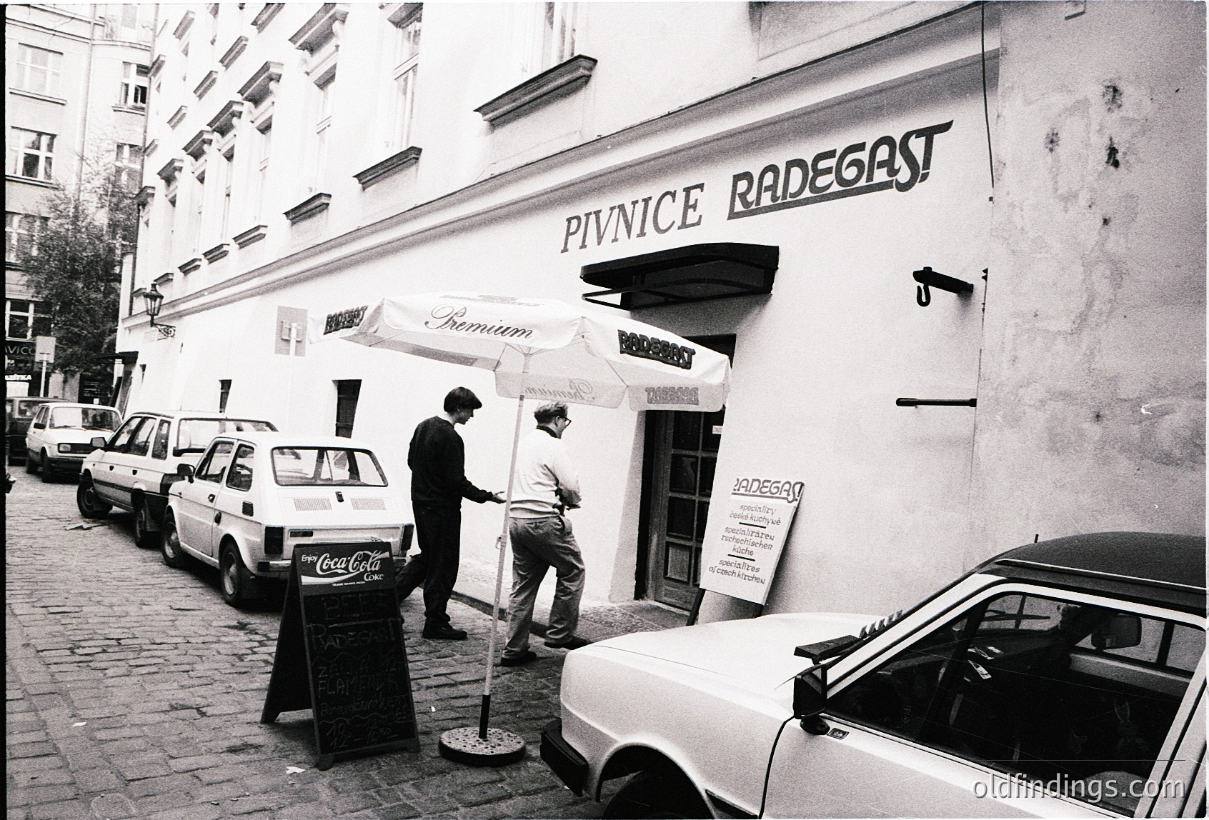 Black-and-white street scene featuring **Pivnice Radegast**, a Czech beer hall, with vintage cars parked along cobblestone pavement. Two men in business attire stand near an open umbrella branded "Premium Radegast." A Coca-Cola sign and signage in Czech script (#český) suggest 1980s–1990s Prague.