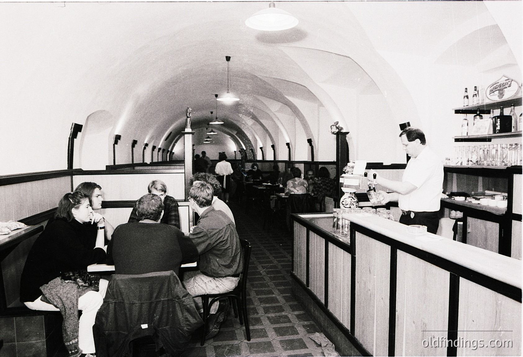 Vintage underground café with arched brick ceilings and tiled floors, featuring 1970s-style decor. Patrons seated at high tables, staff serving drinks behind a counter with shelves displaying bottles. Warm, intimate atmosphere. é