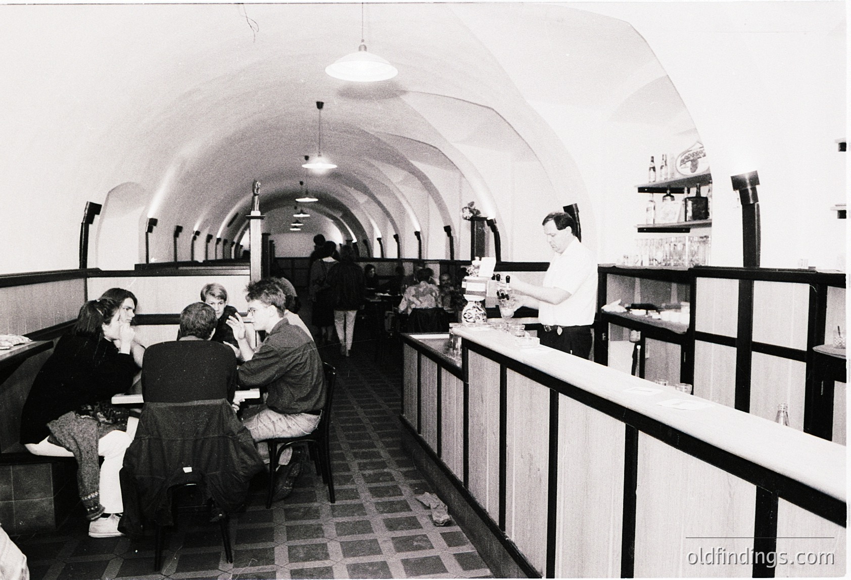 Vintage underground café with curved, arched ceiling and checkered floor. Mid-20th century interior features curved benches, a countertop bar, and patrons in 1960s-era clothing. Industrial lighting and exposed pipes add to the retro aesthetic. é