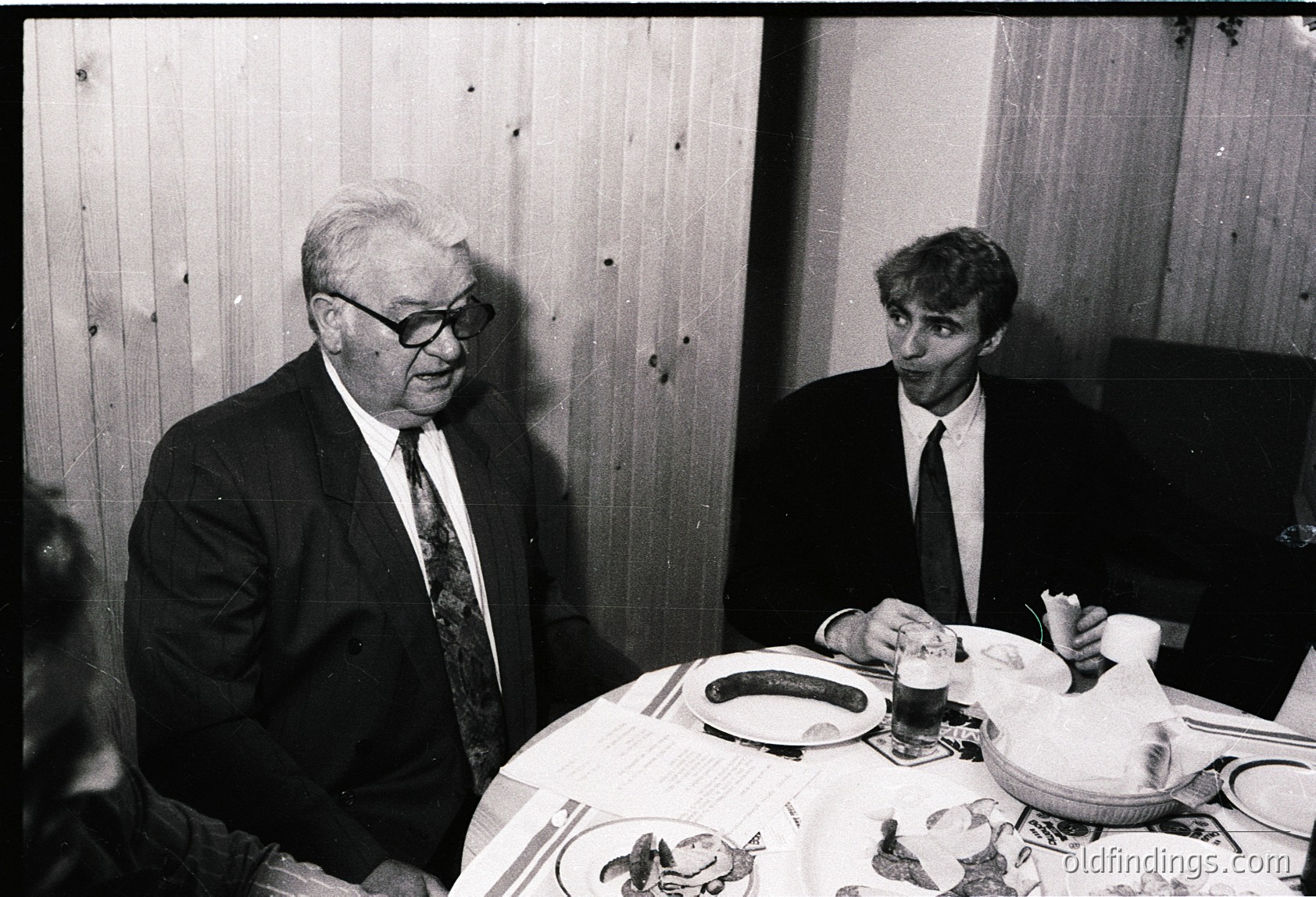 Two men in formal attire share a meal at a wooden-paneled table, likely mid-20th century. The older man wears glasses and a suit, while the younger man holds a fork. A menu and empty plates suggest a restaurant or formal dining setting.