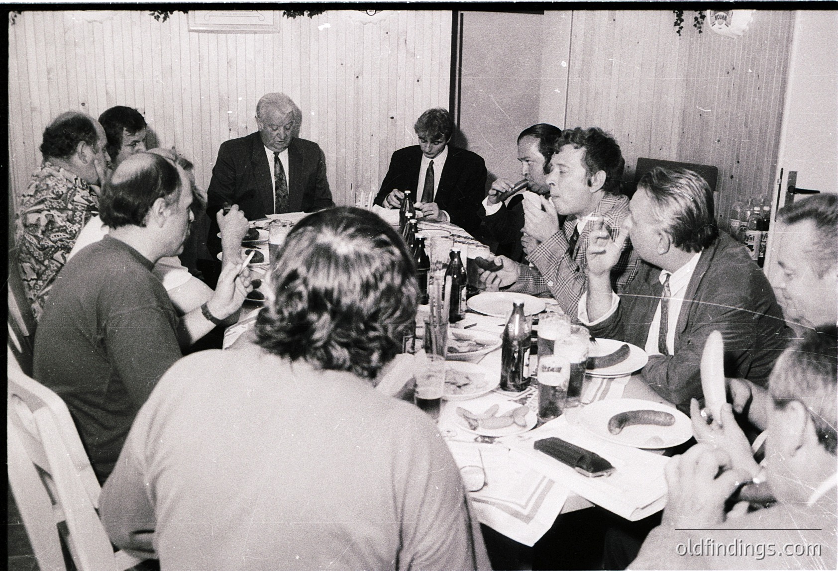 Group of 15+ men and women seated at a long table in a formal indoor setting, likely a 1970s–1980s business or diplomatic gathering. White tablecloth, paper plates, and disposable cutlery suggest a mid-century event. One man in a suit stands at the head, gesturing. Bottles of beer and glasses visible. Plain walls and fluorescent lighting indicate a functional venue.