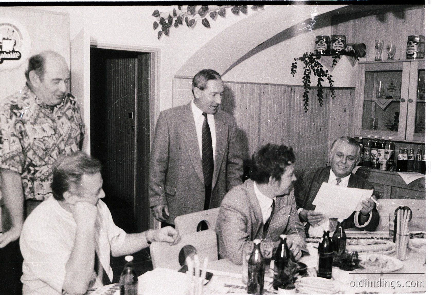 Mid-century indoor gathering in a modest kitchen/dining area, featuring five adults seated/standing around a table. Men wear suits with ties, women in blouses; one man holds a document. Bottles, glasses, and a cake suggest a celebratory meal. Decor includes artificial greenery and a framed poster. Likely Eastern Bloc era (1960s–1980s), possibly Bulgaria or similar region.