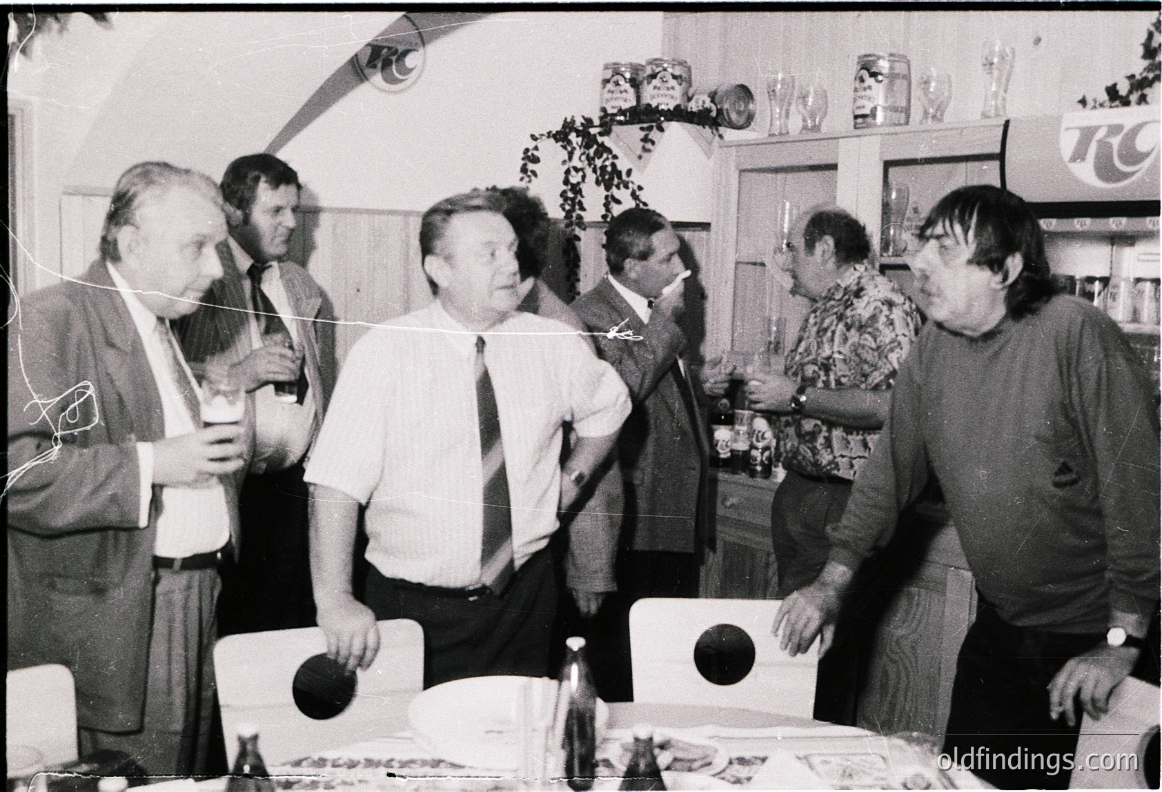 Group of six men in a 1970s-era indoor setting, likely a social gathering. Centered around a table with food, drinks (bottles, glasses), and a cake. Decor includes a "TC" sign and floral wall art. Formal and casual attire suggests mixed professional/relaxed atmosphere.