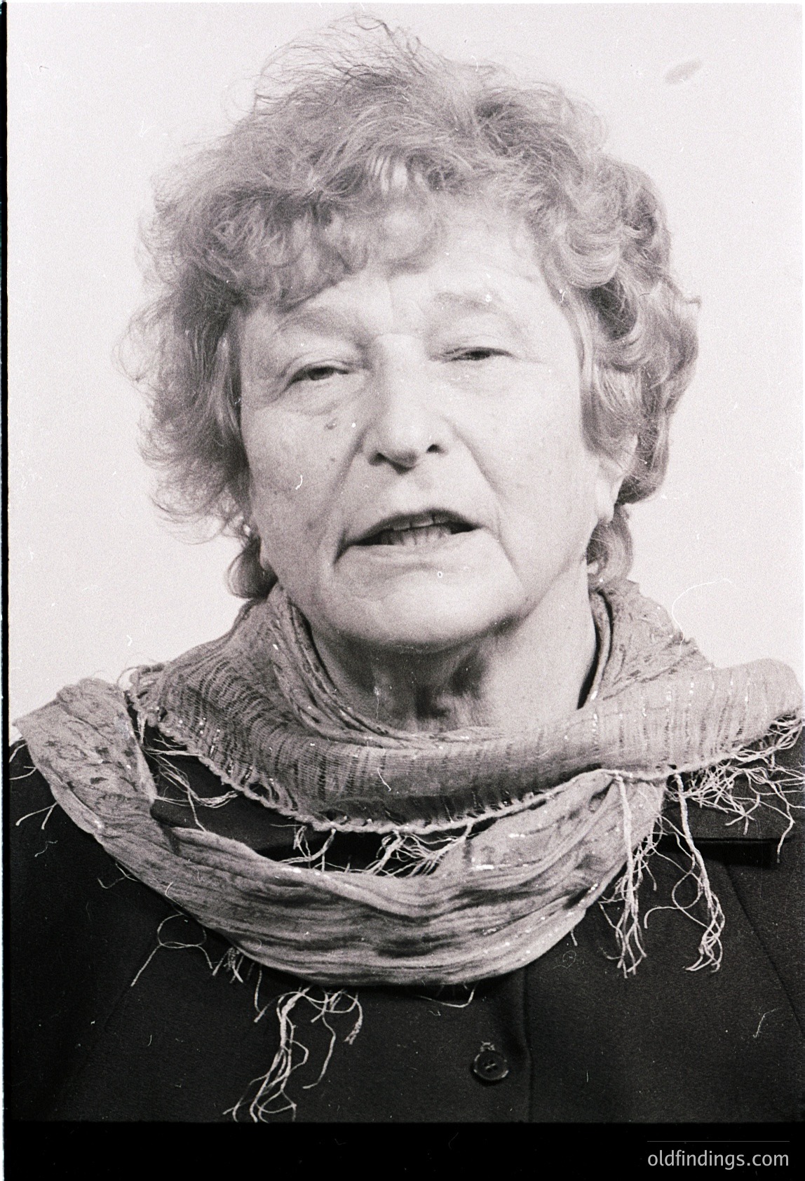 Portrait of an elderly woman in mid-20th century attire, likely –. She wears a dark jacket with a frayed, textured scarf draped over her shoulders. Expression suggests contemplation or seriousness. Style hints at European or North American mid-century fashion.